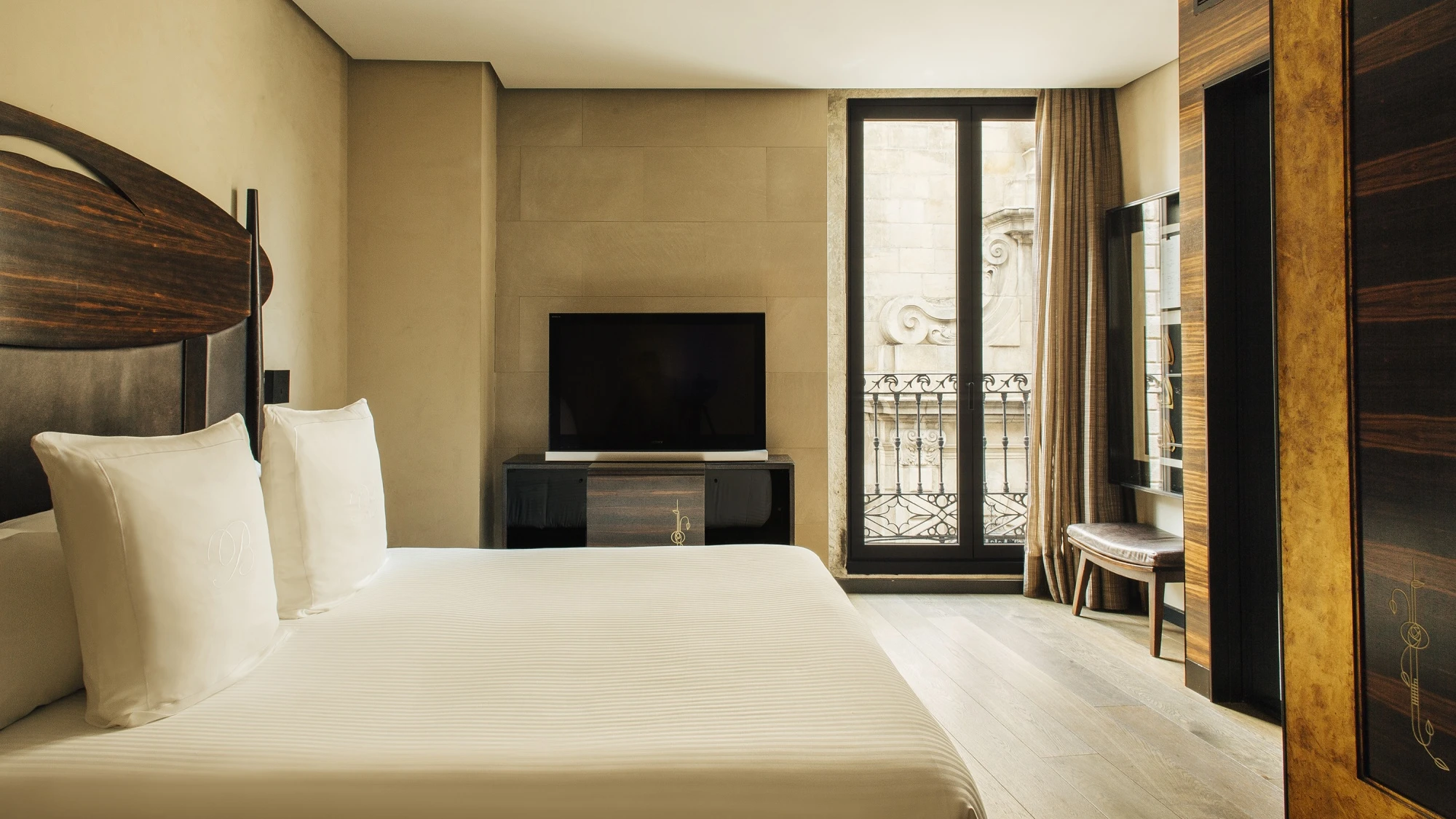 Se ve una cama con ropa de cama clara, una televisión y una puerta de balcón con cortinas en una habitación luminosa.