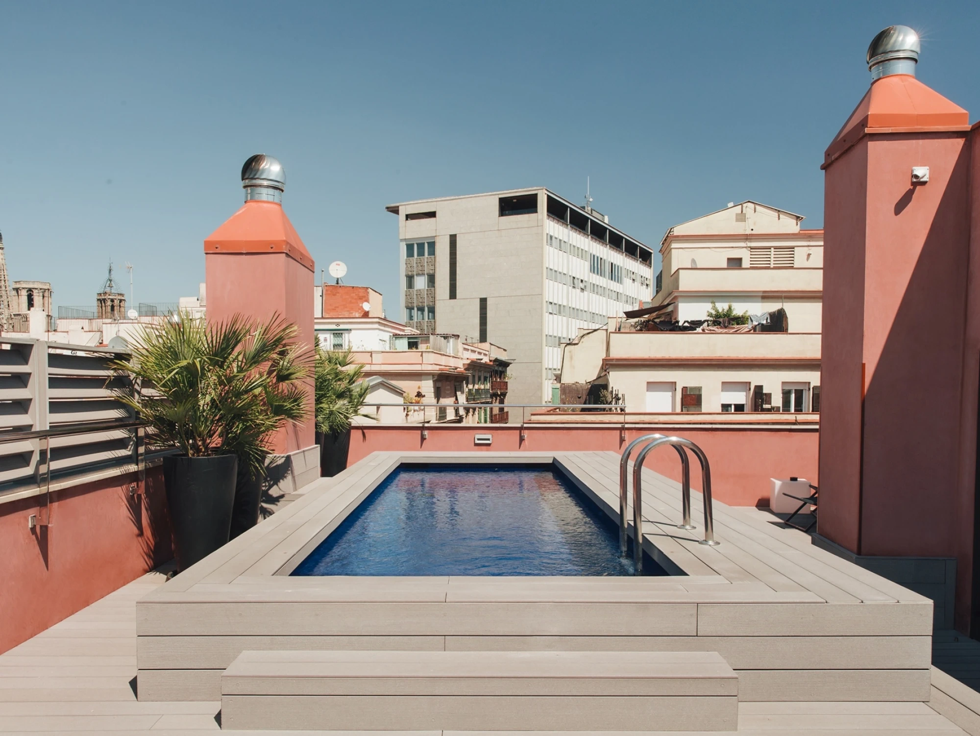 Se ve una piscina rectangular al aire libre con paredes de color rosa y plantas en macetas alrededor.
