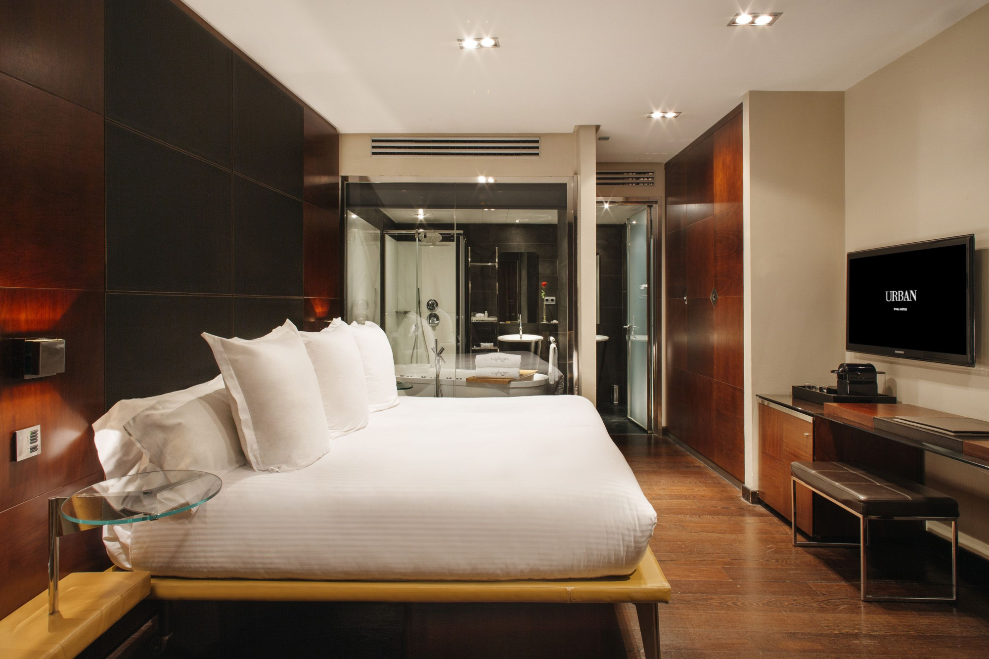 Se ve una cama con ropa de cama blanca, un televisor y un escritorio en una habitación con suelo de madera.