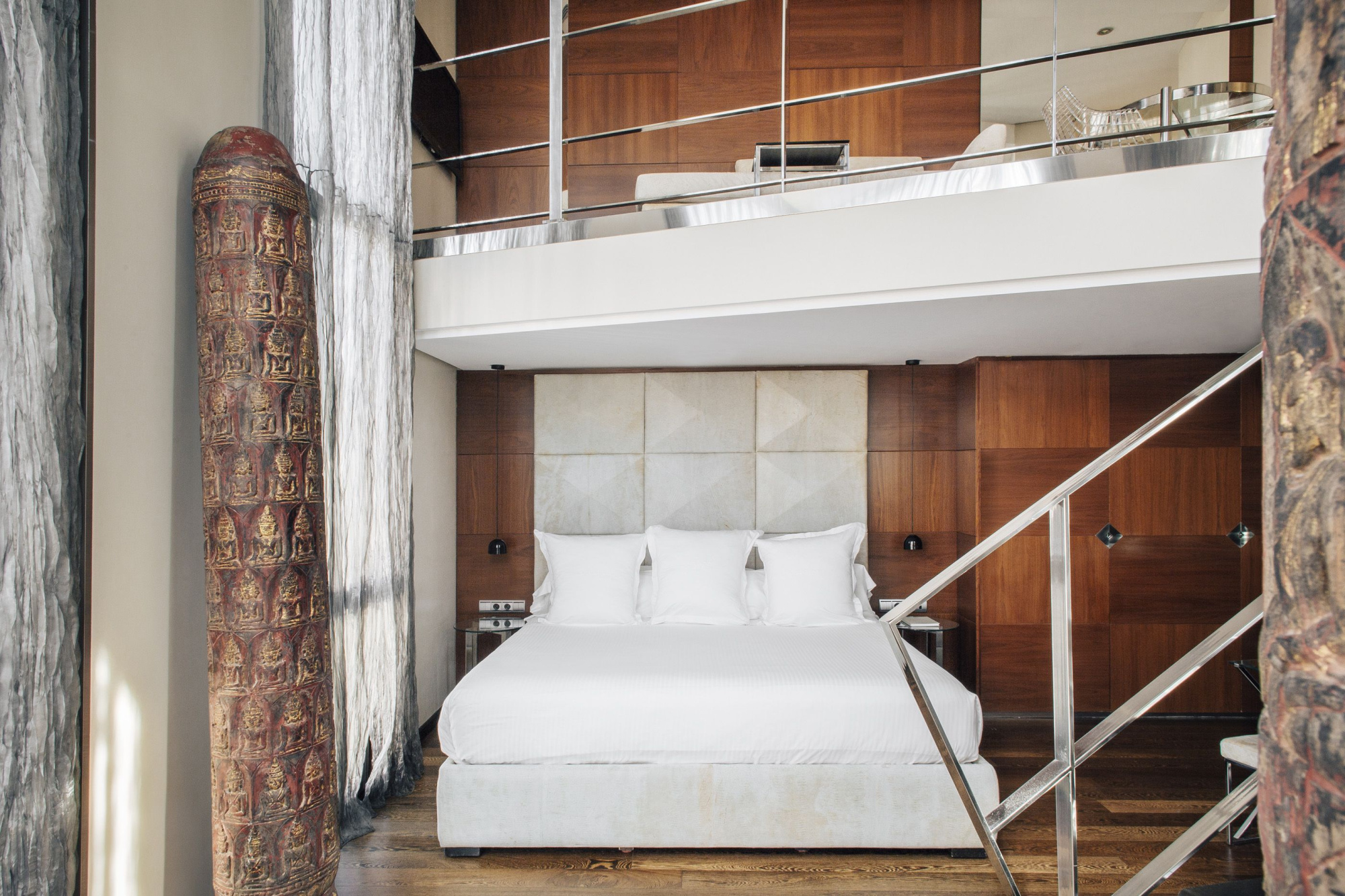Se ve una cama doble con ropa de cama blanca y una zona de estar junto a una escalera de metal.