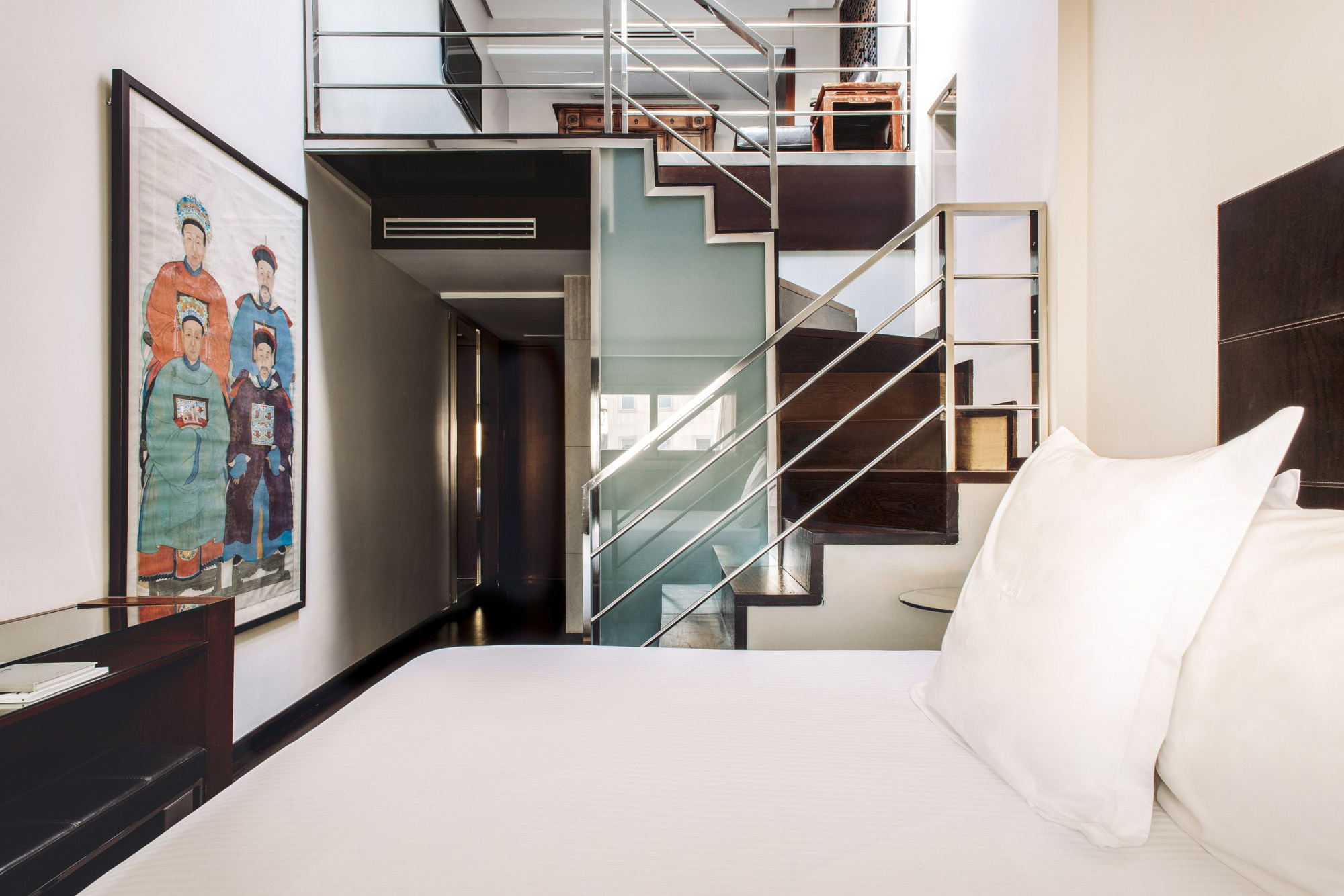 Se ve una cama con ropa de cama blanca y una escalera interior de metal y vidrio al fondo.