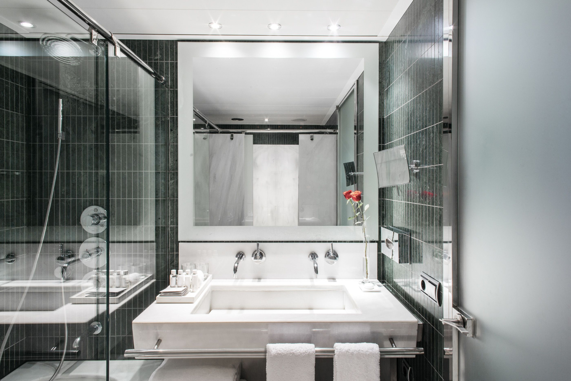 Se ve un baño moderno con ducha de cristal, dos lavabos y toallas dobladas sobre el mueble.