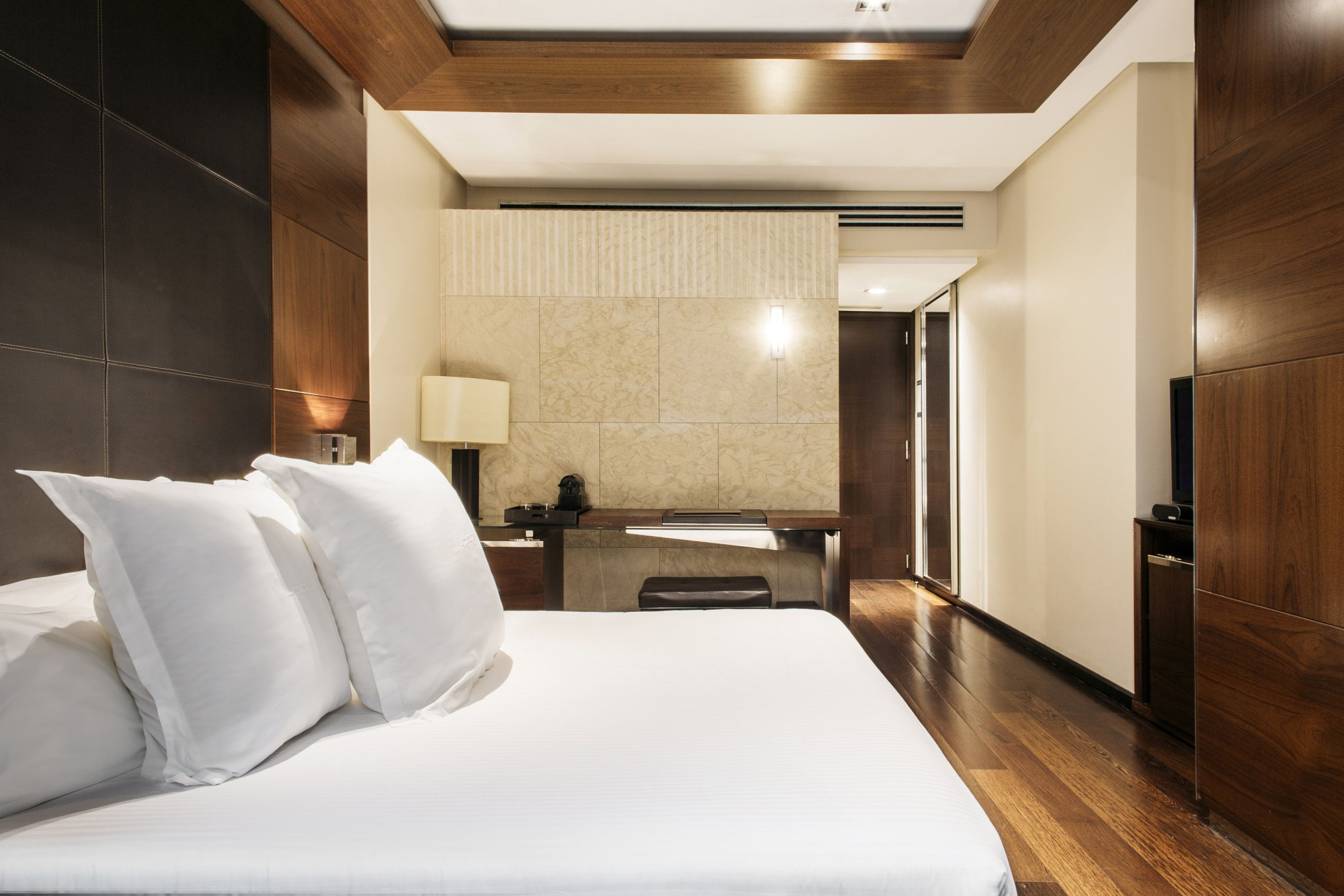 Se ve una cama con cojines blancos y una zona de estar al fondo con sofá y suelo de madera.