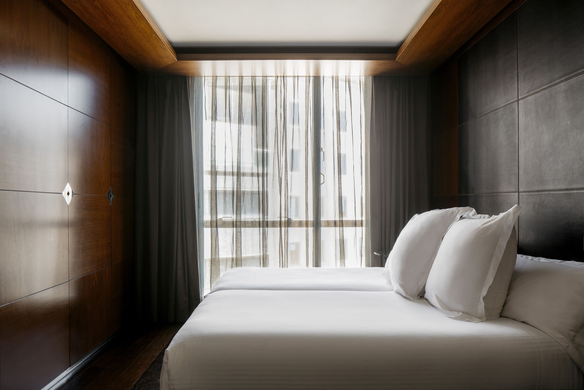Se ve una cama doble con almohadas y cortinas frente a una ventana, en una habitación con paredes de madera y azulejos grises.