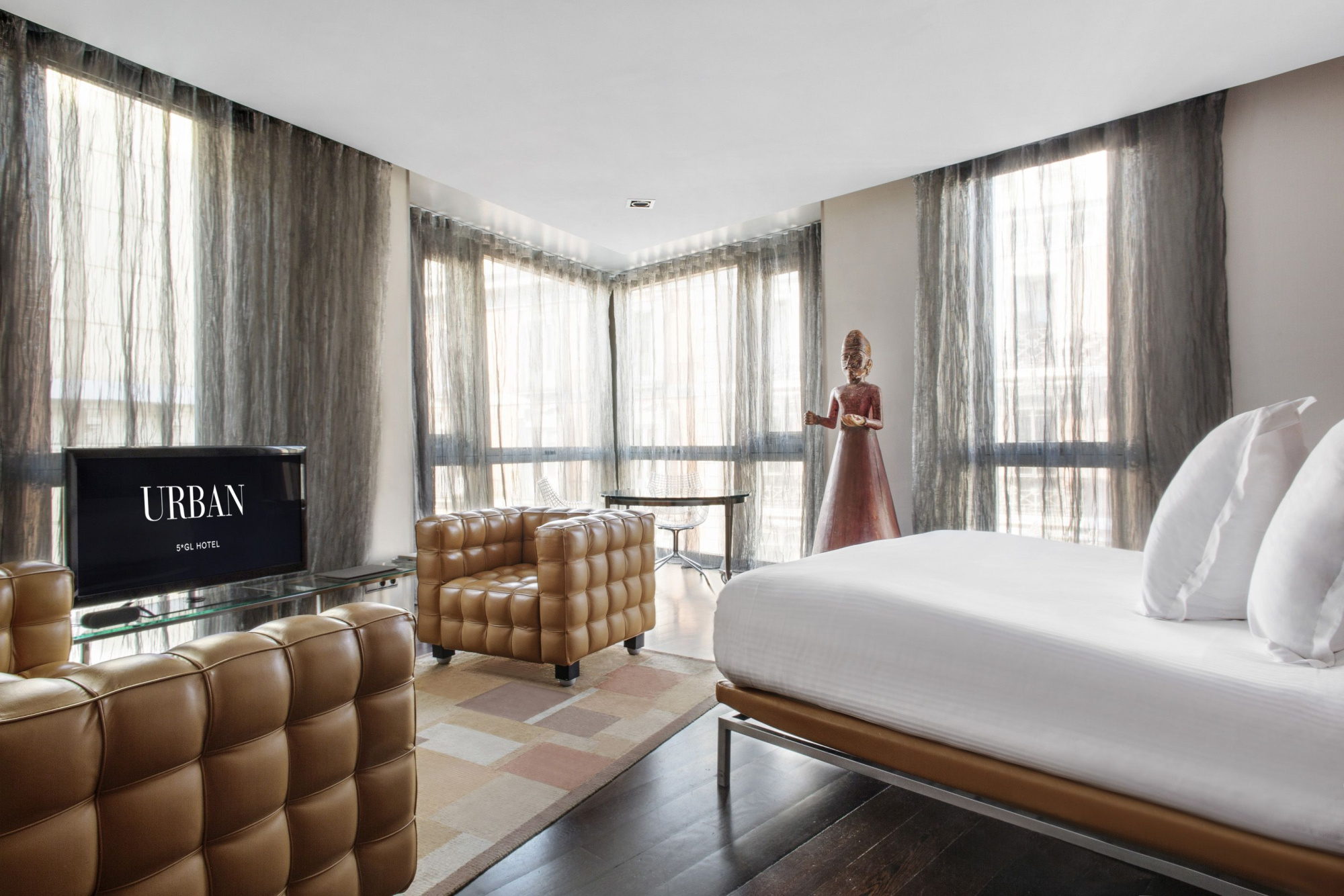 Se ve una habitación con una cama doble, sillones de cuero y cortinas claras junto a una ventana grande.