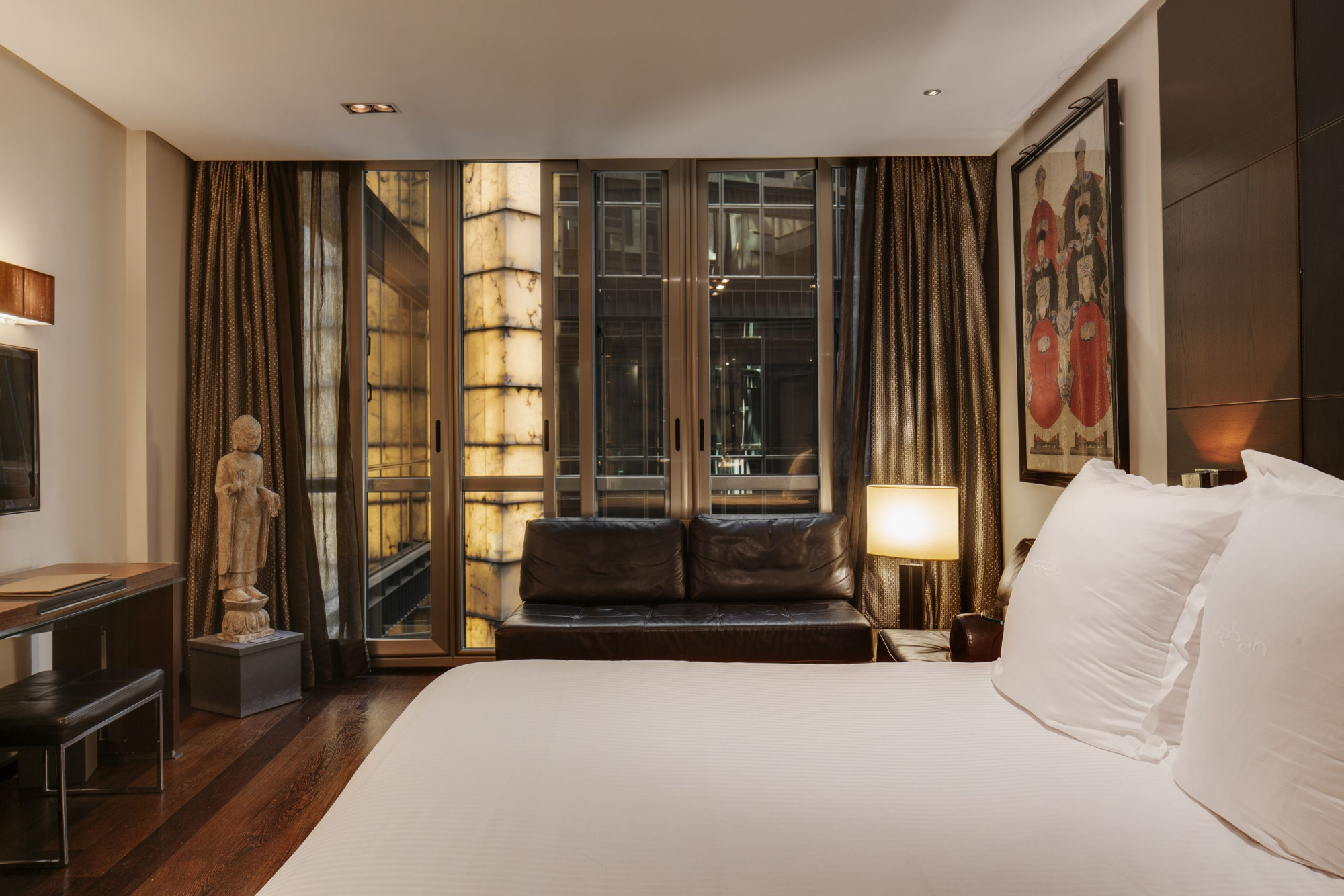 Se ve una cama con ropa blanca, un sillón oscuro y un gran ventanal con cortinas en una habitación.