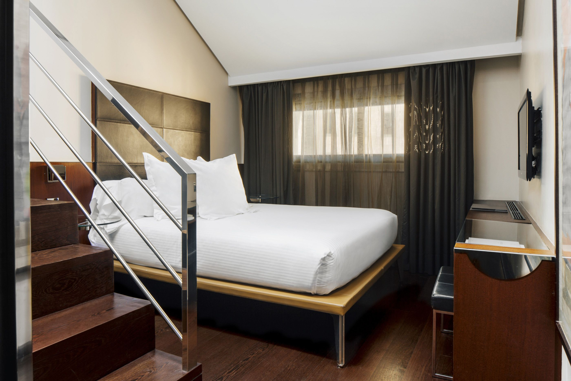 Se ve una cama doble con ropa de cama blanca, cortinas oscuras y una escalera metálica junto a una zona de estar.