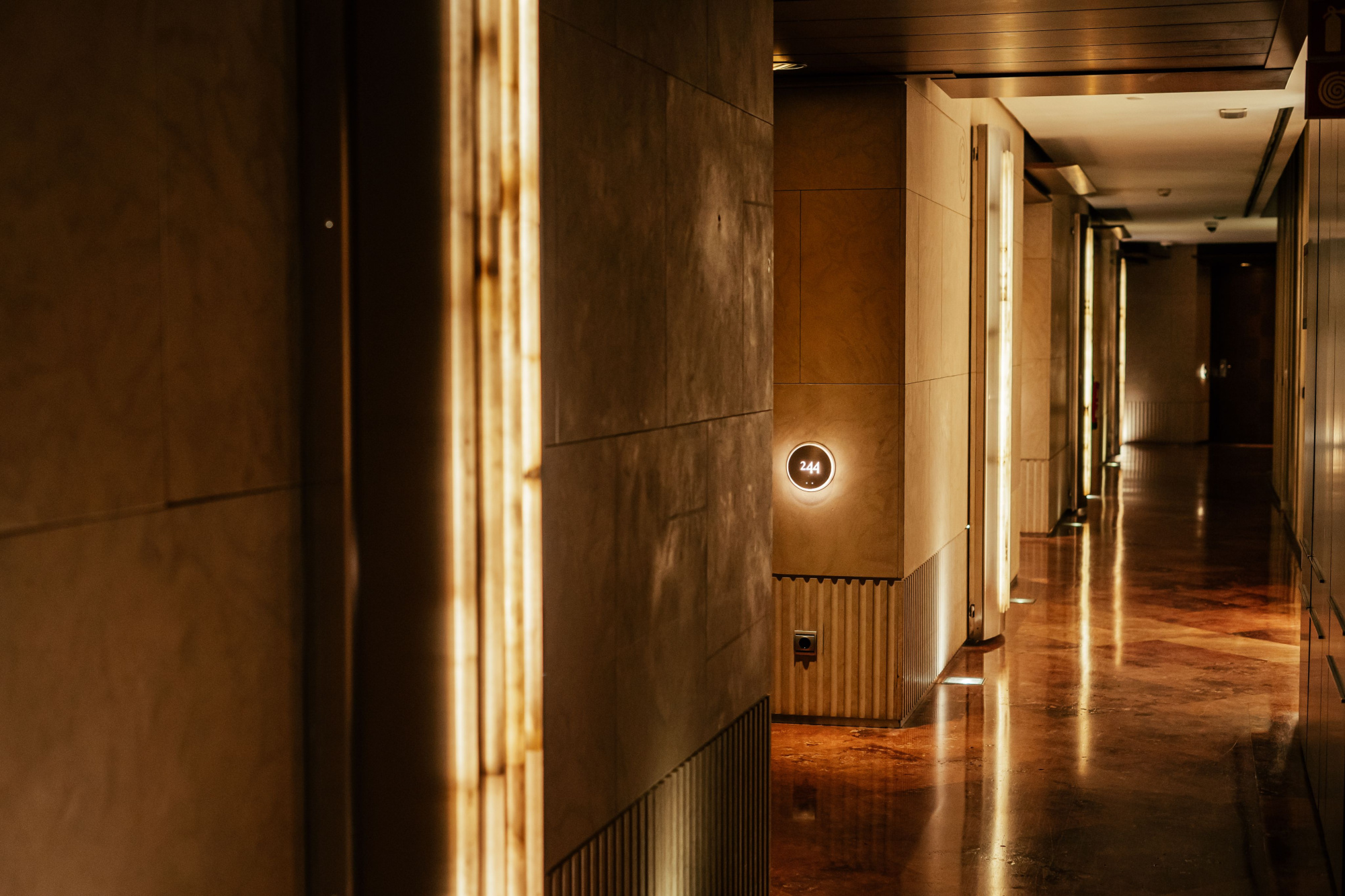 Pasillo interior con paredes de ladrillo visto y suelo de madera, iluminado con luces cálidas empotradas.