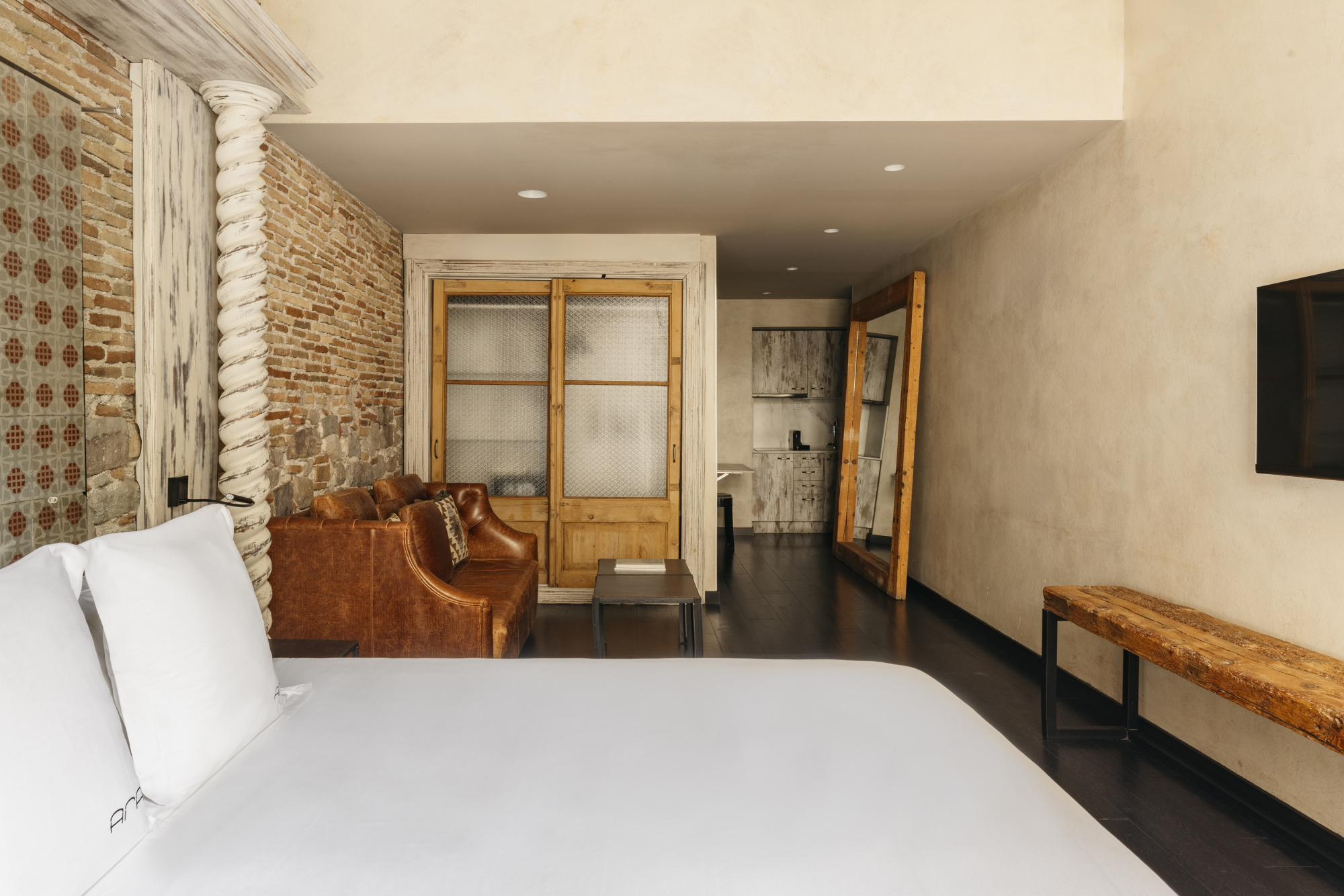 Se ve una cama con ropa de cama blanca, un sillón de cuero y una zona de estar con TV en una habitación.