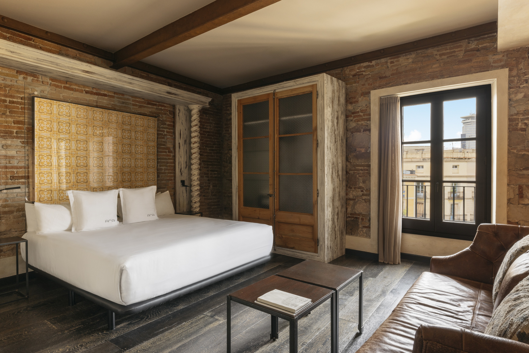 Se ve una cama doble con ropa de cama blanca, un sillón marrón y una ventana con luz natural en la habitación.