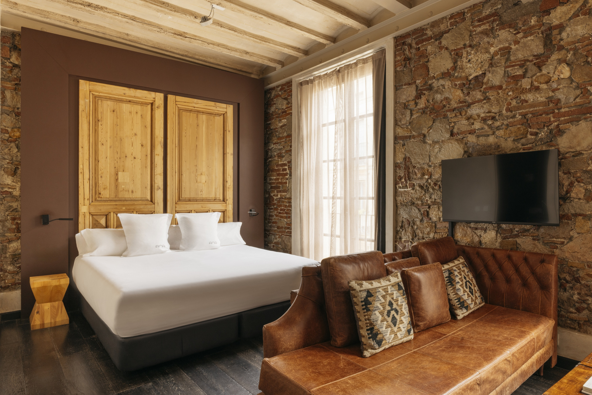 Se ve una cama doble, un sillón de cuero y una pared de ladrillo con una televisión y una ventana con cortinas.