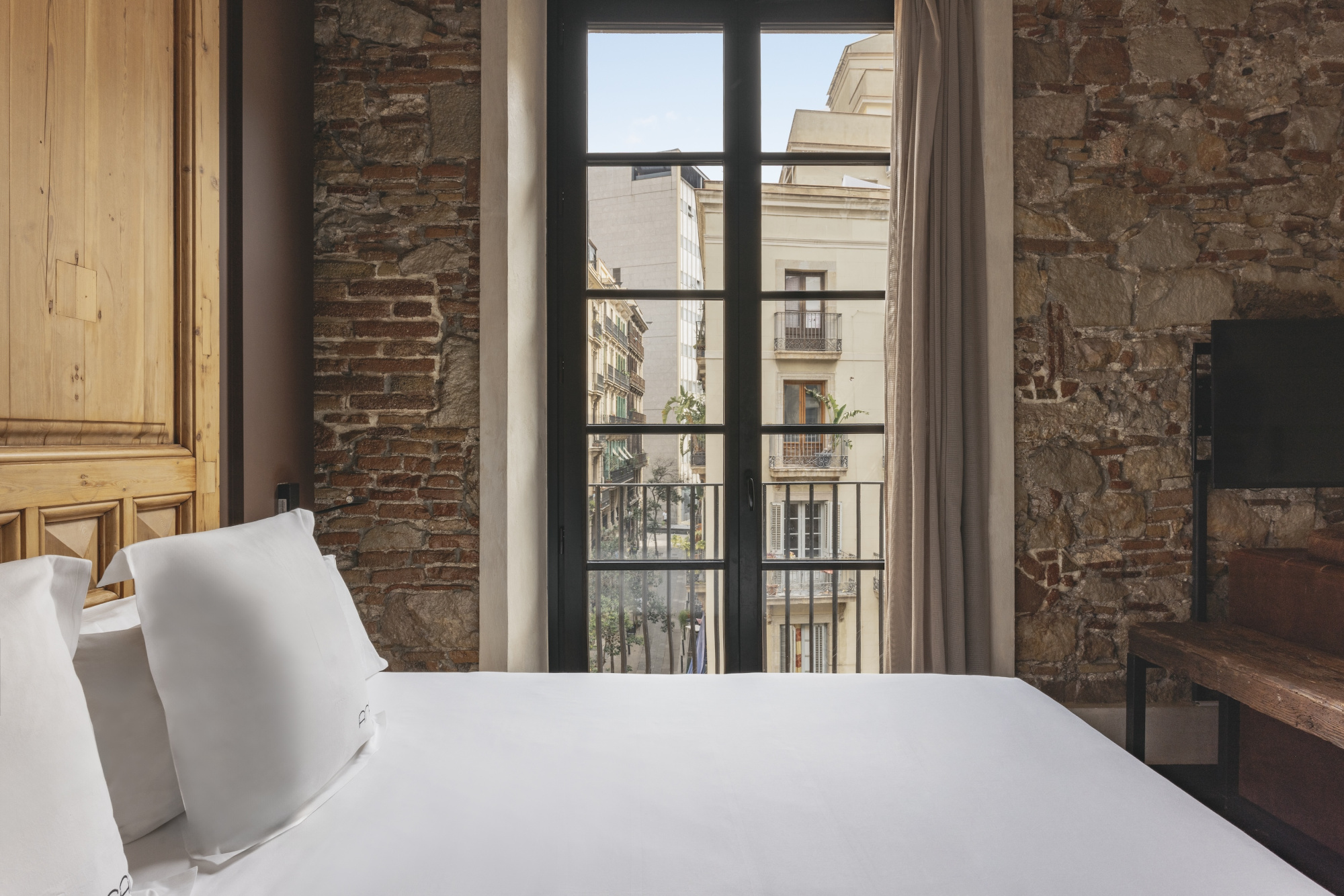 Se ve una cama con ropa blanca y cojines junto a una ventana con barandilla de balcón al fondo.