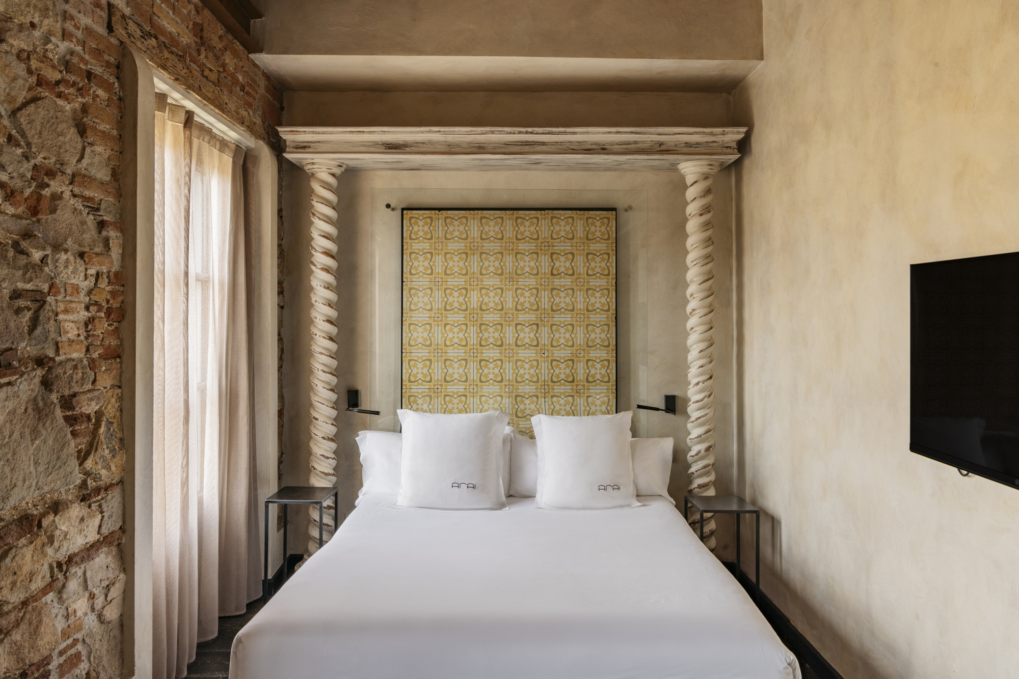 Se ve una cama doble con almohadas y una pared con cabecero textil, junto a cortinas y una ventana lateral.