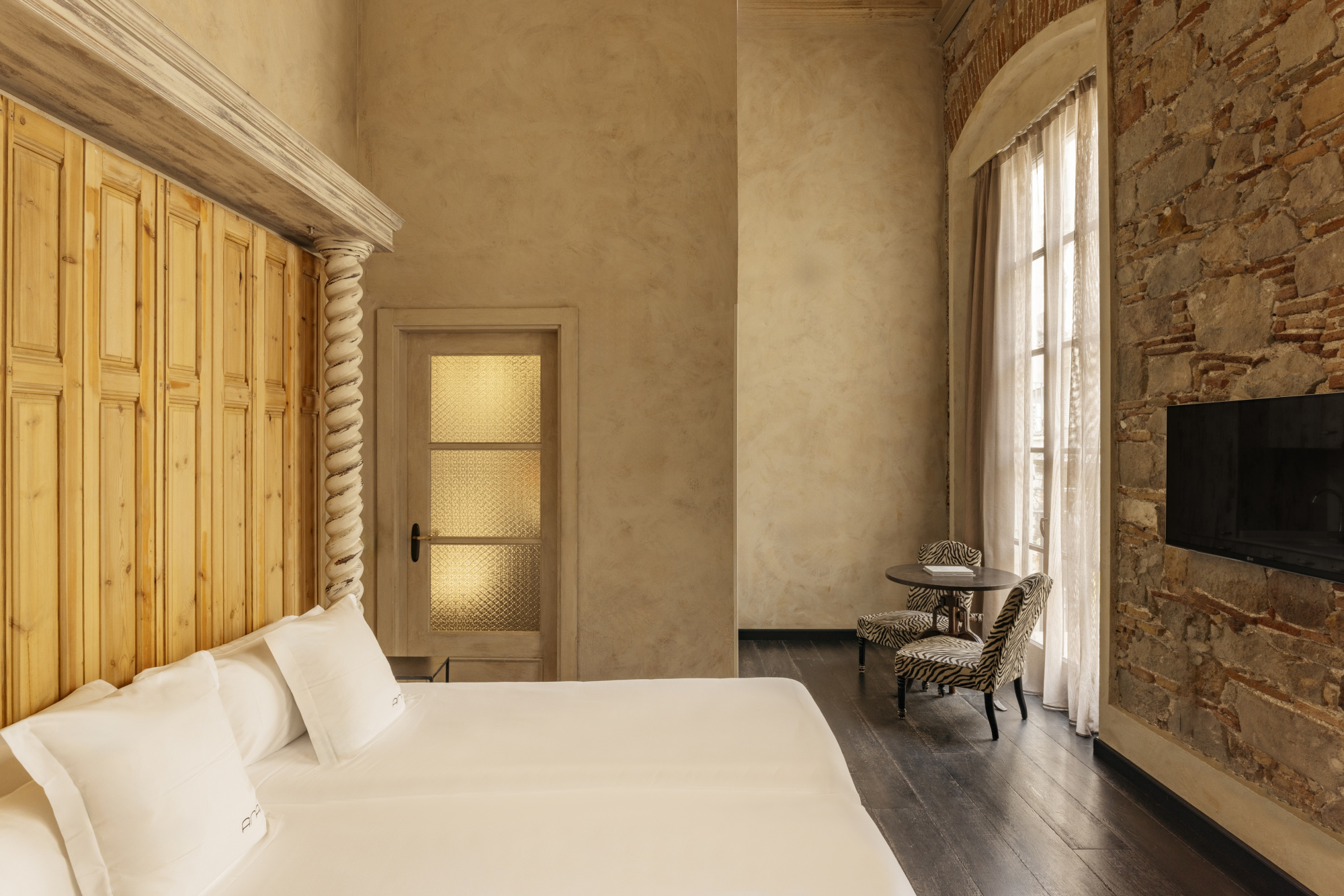 Se ve una cama con ropa de cama blanca, una pared de piedra y una ventana junto a una silla y una mesa auxiliar.