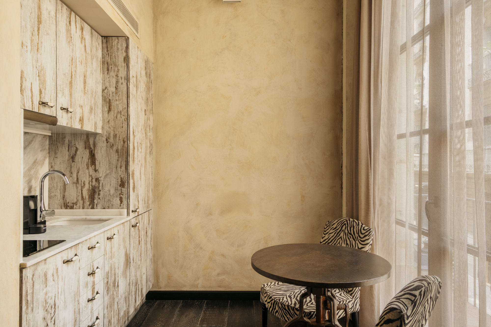 Se ve un rincón interior con una mesa redonda, dos sillas y cortinas junto a una pared beige.