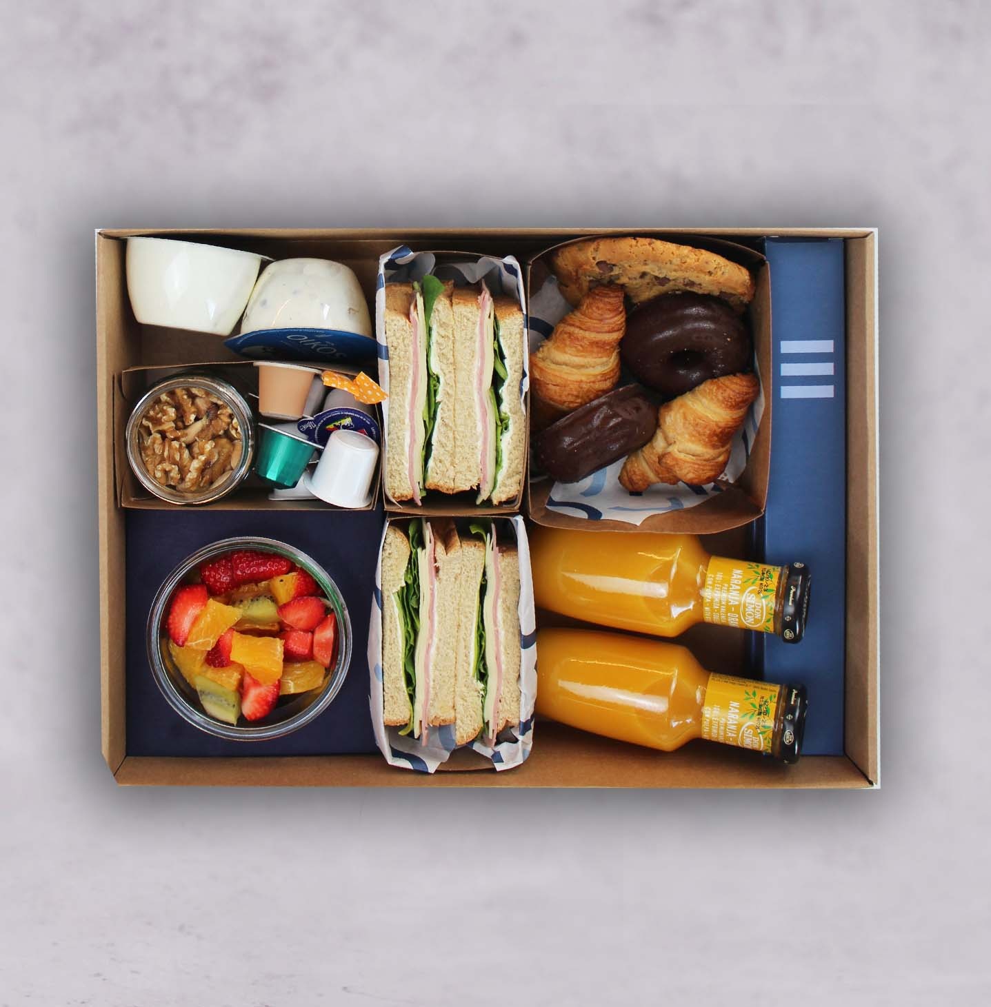 Se ve una caja de desayuno con croissants, sándwiches, fruta, yogur y botellas.