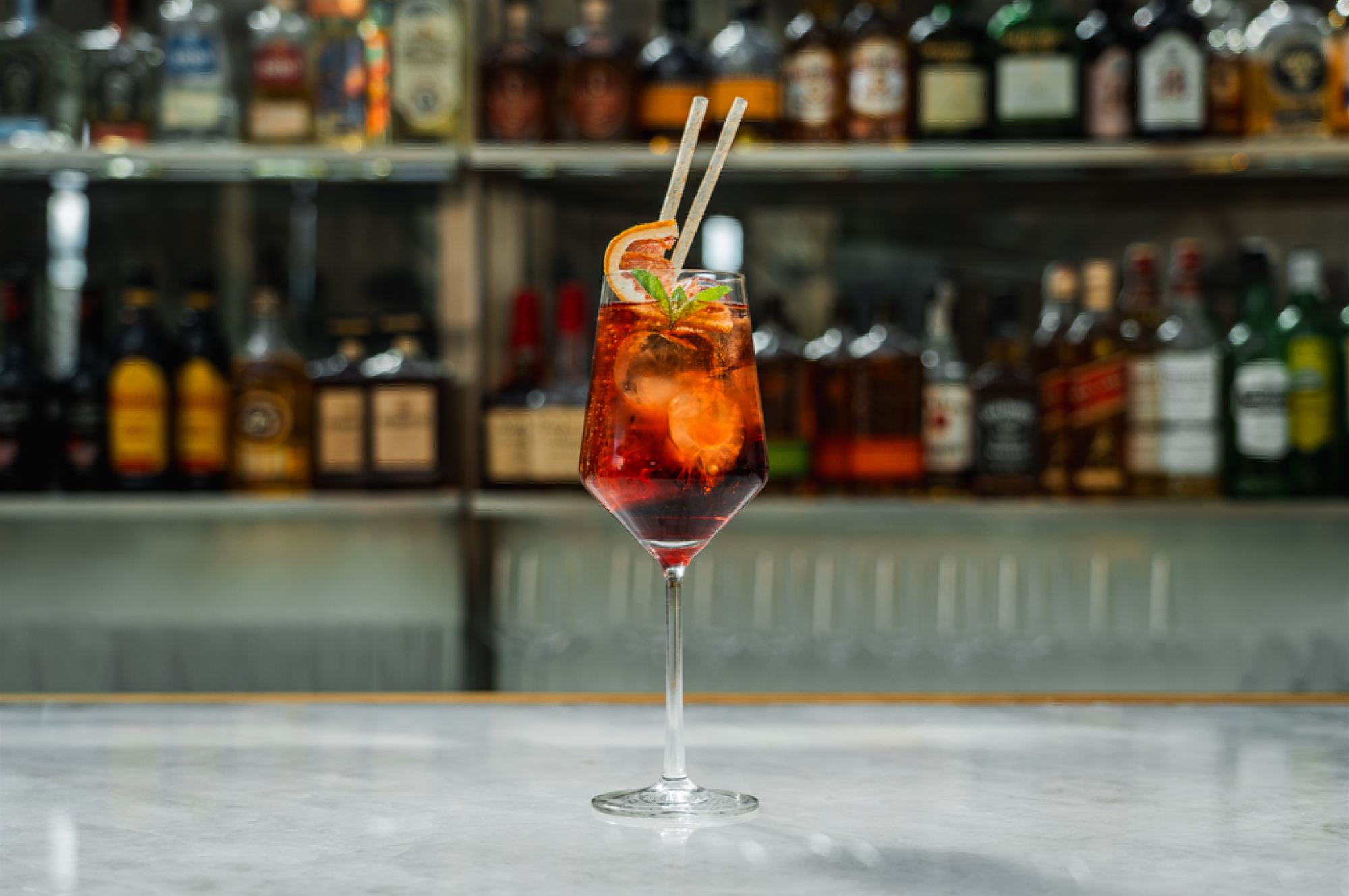 Sirve un cóctel con hielo y pajitas sobre una barra con botellas al fondo.