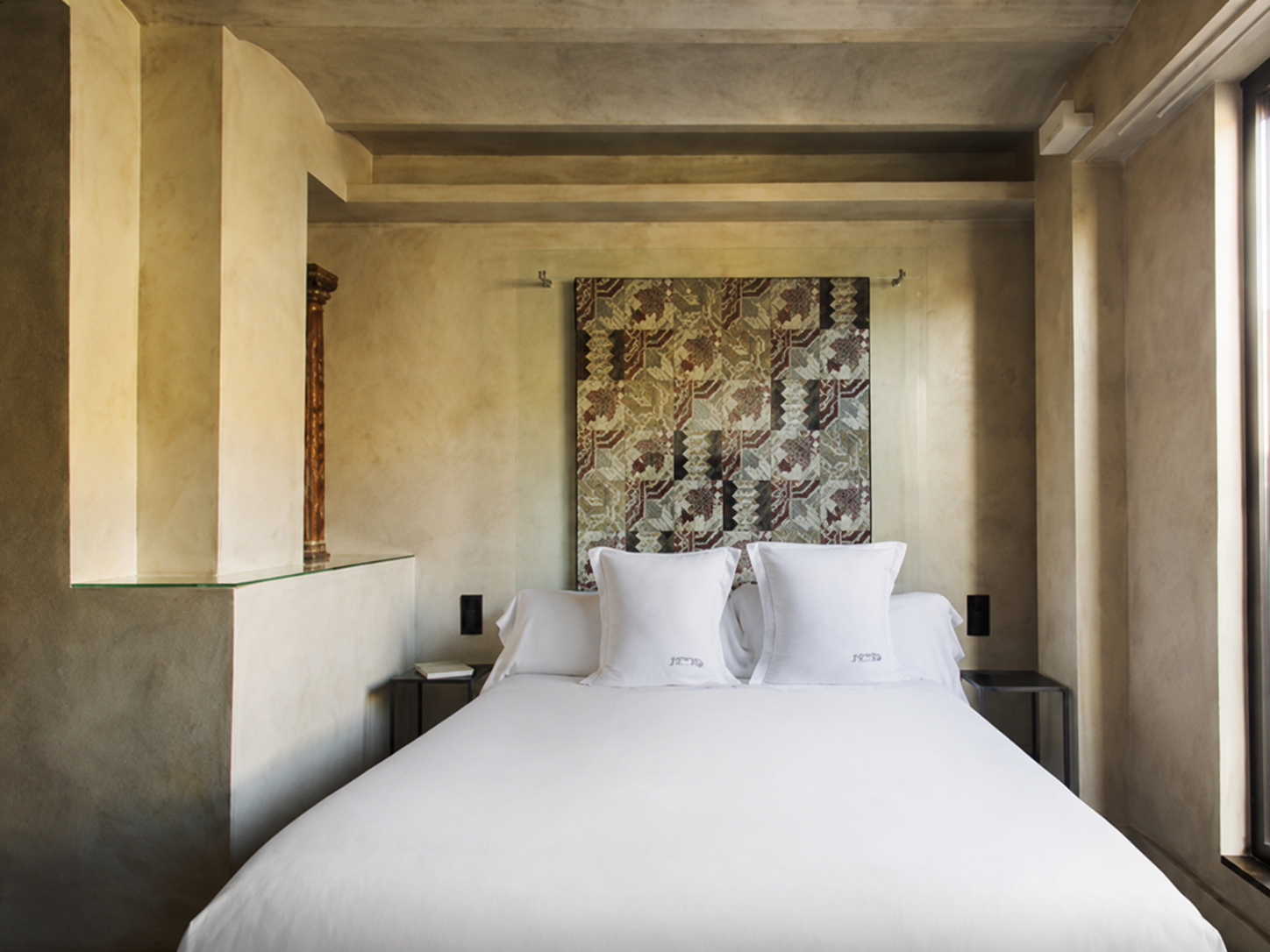 Se ve una cama doble con ropa de cama blanca y dos almohadas, junto a paredes claras y una ventana lateral.