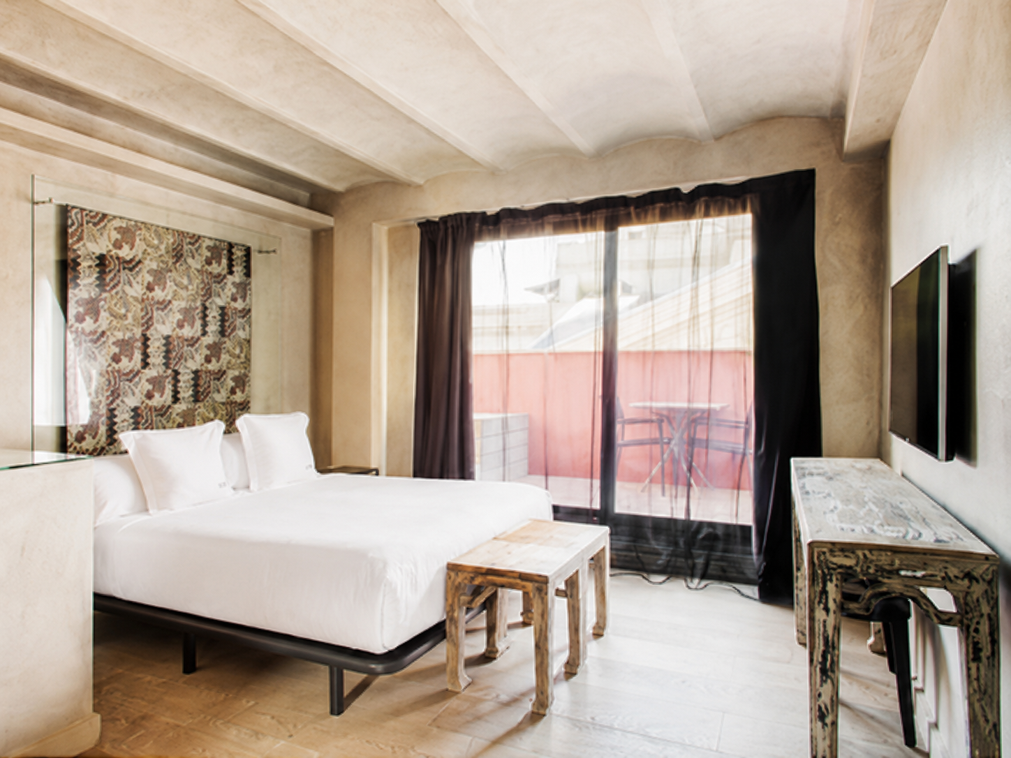 Se ve una cama doble con ropa de cama blanca, una mesa auxiliar de madera y una puerta con cortinas y acceso a un balcón.