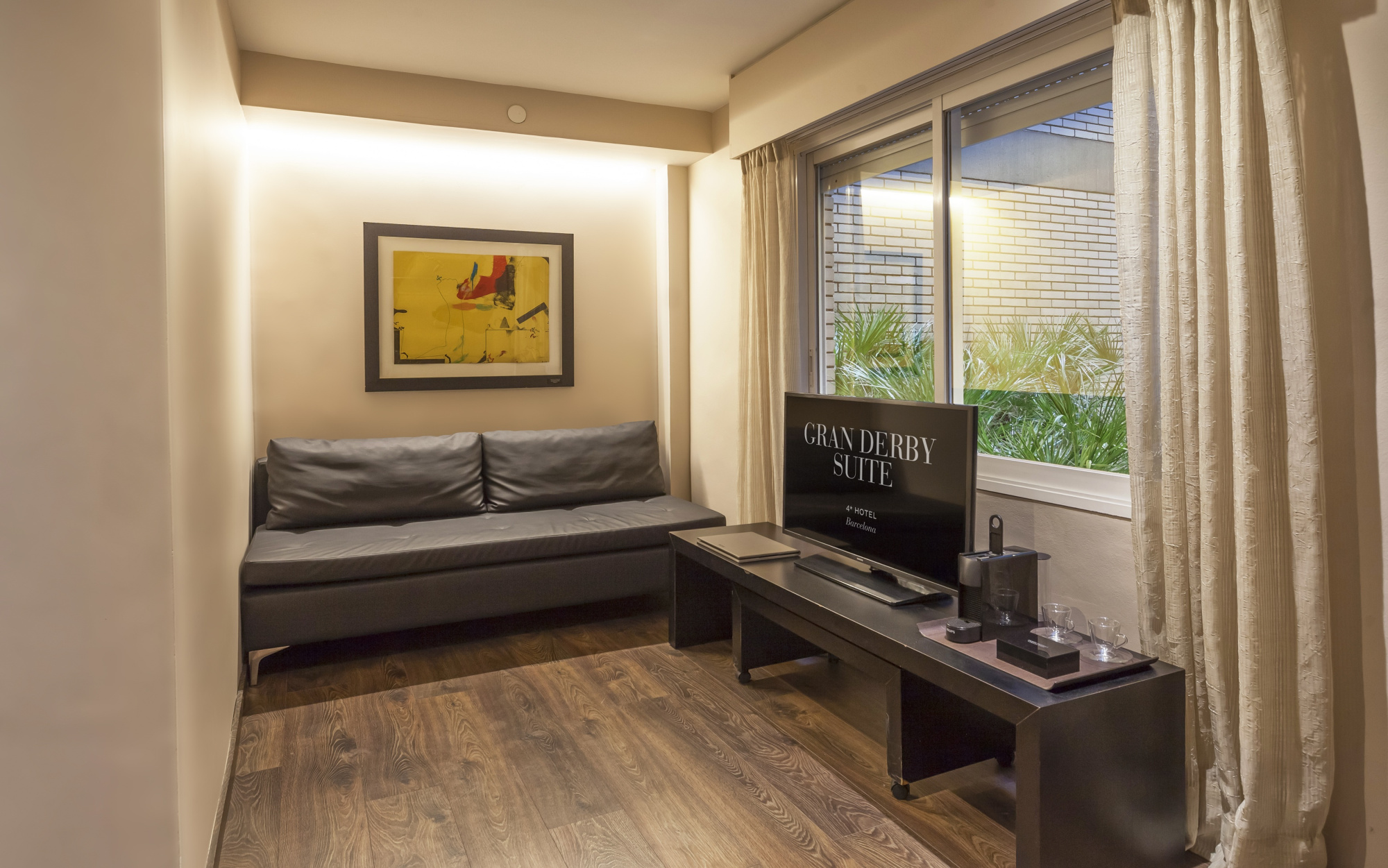 Se ve una cama, un escritorio con silla y una ventana con cortinas en una habitación luminosa.