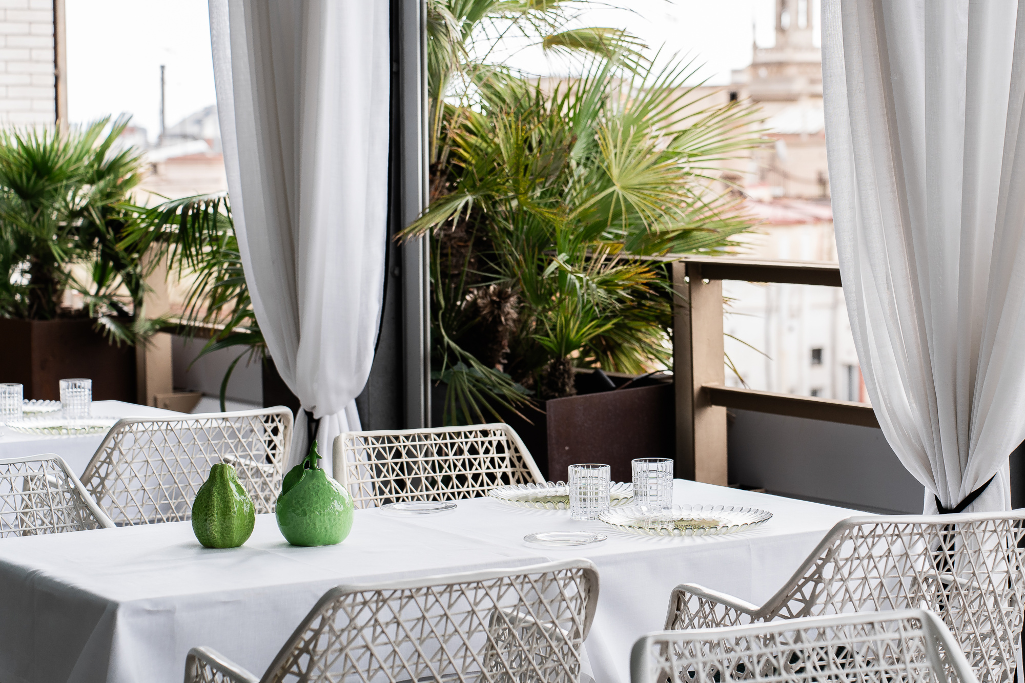 Se ve una mesa con mantel blanco y sillas de rejilla en una terraza con plantas y cortinas blancas.