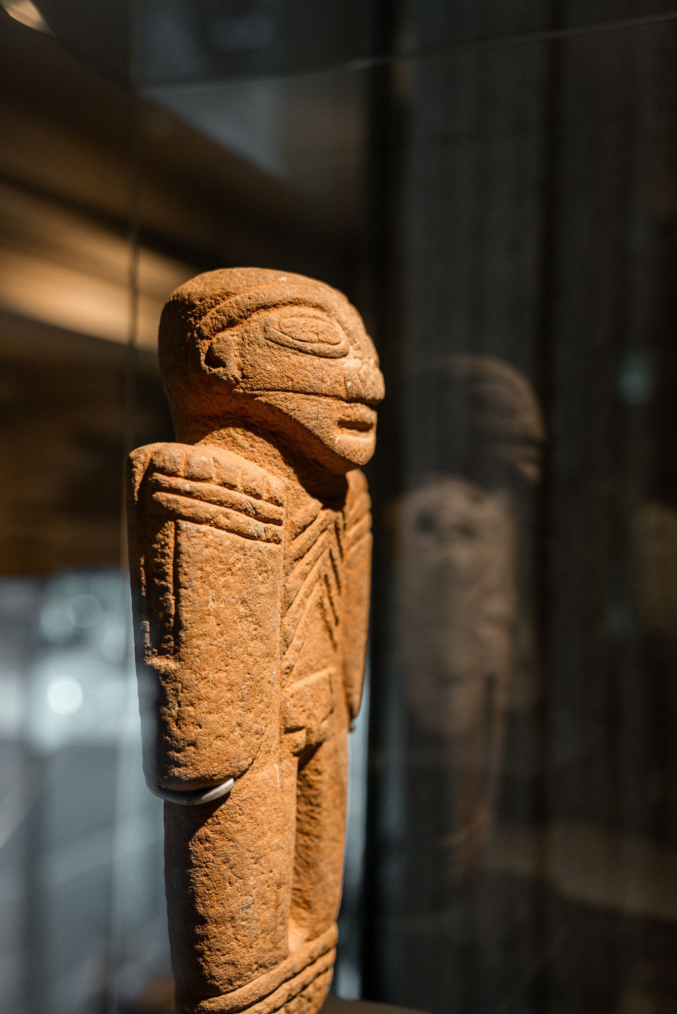 Se ve una escultura de piedra con forma humana sobre un pedestal en un espacio interior con vitrinas al fondo.