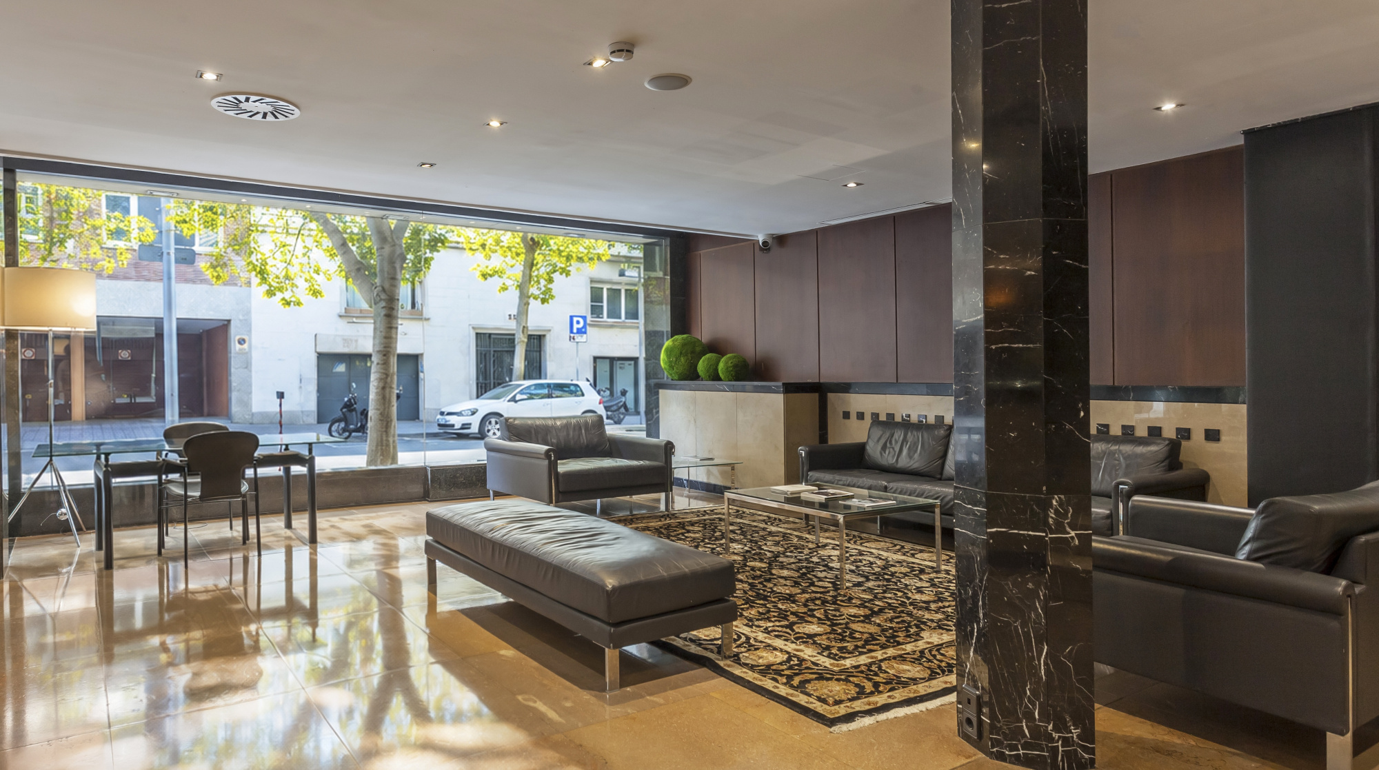 Se ve una zona de estar con sofás, una mesa auxiliar y ventanales que dan a la entrada exterior.