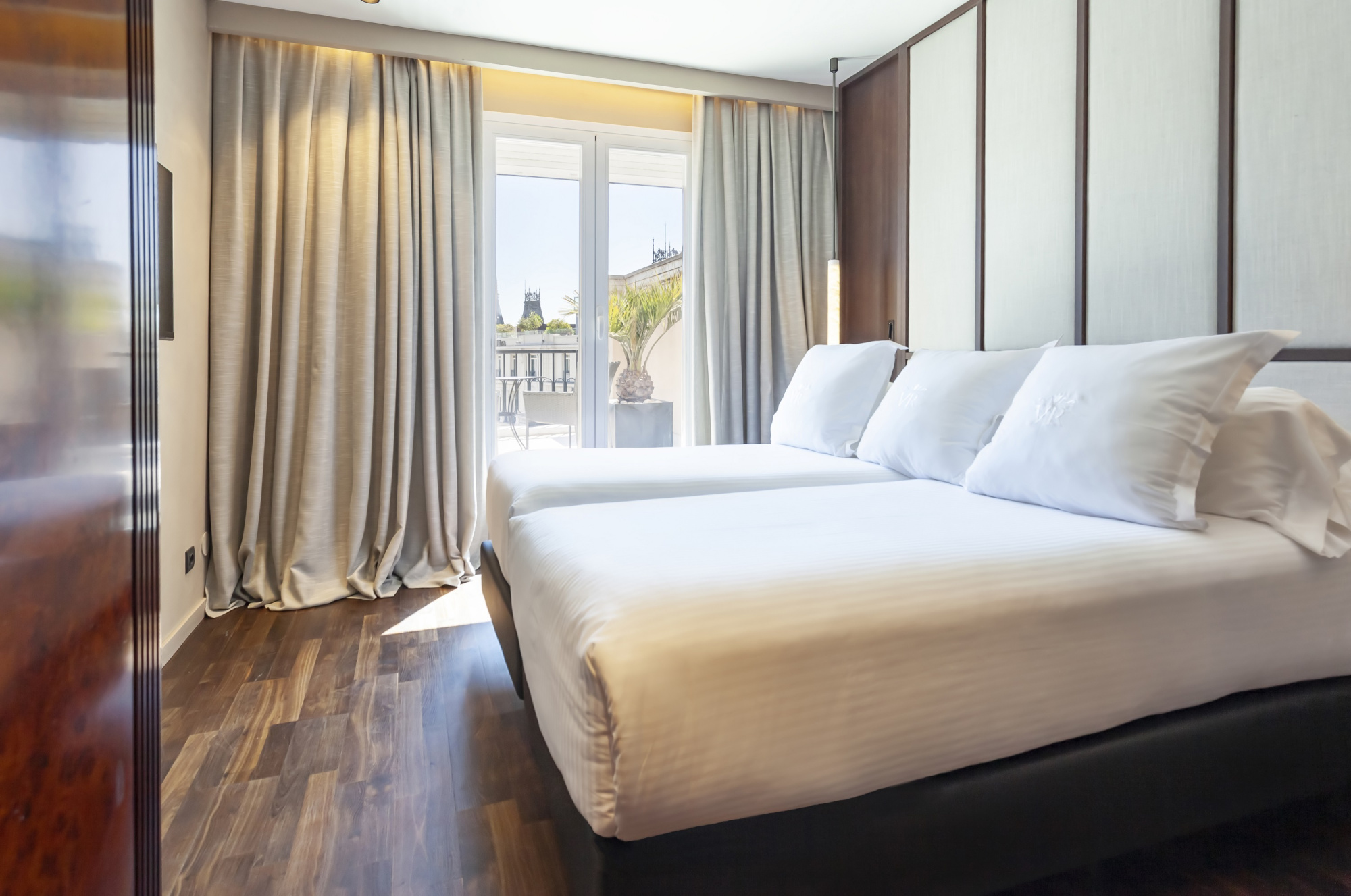 Se ve una cama doble con ropa de cama blanca y un ventanal con cortinas junto a una mesa auxiliar.