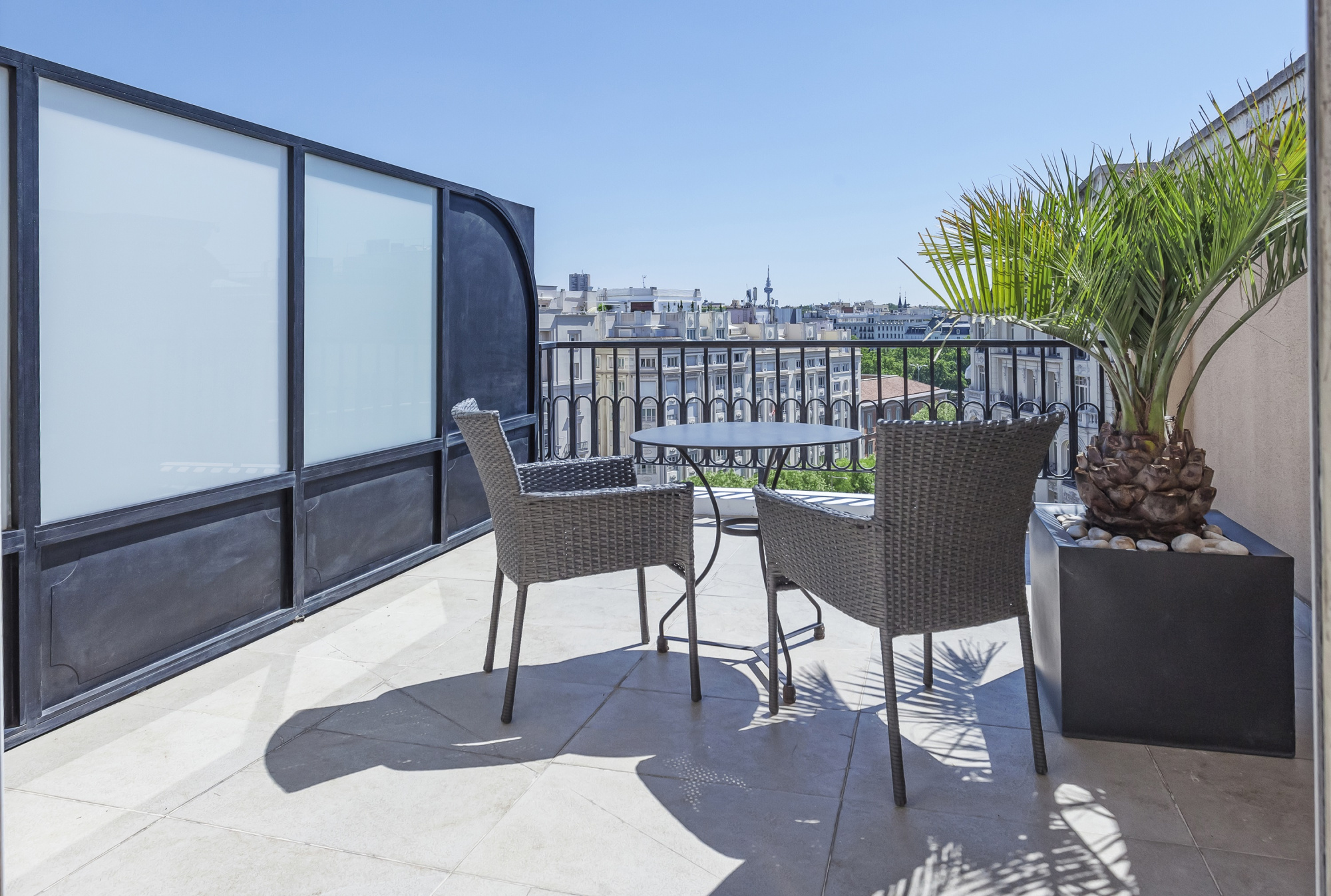 Se ve una terraza soleada con una mesa pequeña, dos sillas y una barandilla de metal junto a una pared blanca.