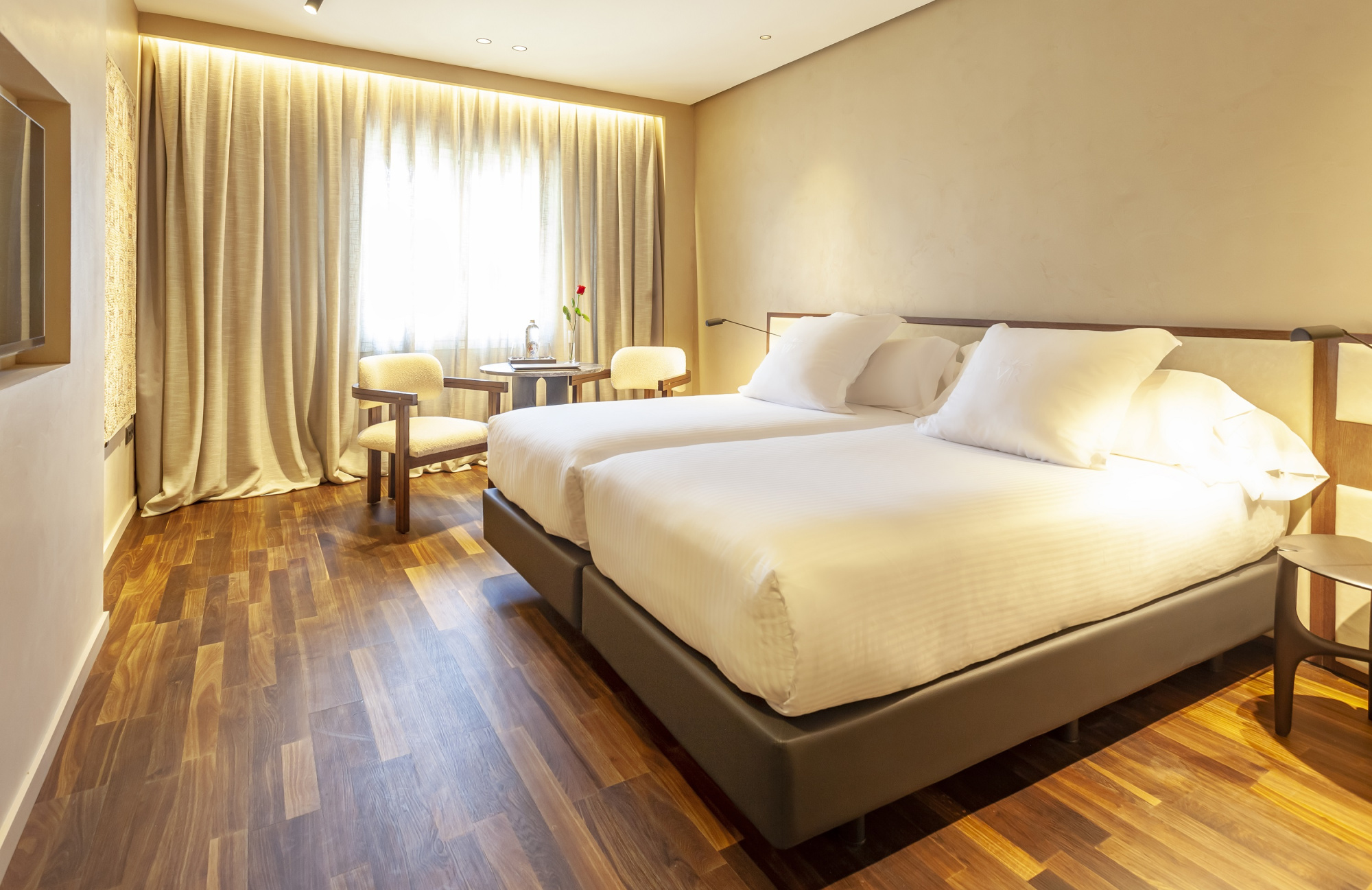 Una cama doble con ropa de cama blanca y cortinas beige se ve en una habitación con suelo de madera y dos sillones junto a la ventana.