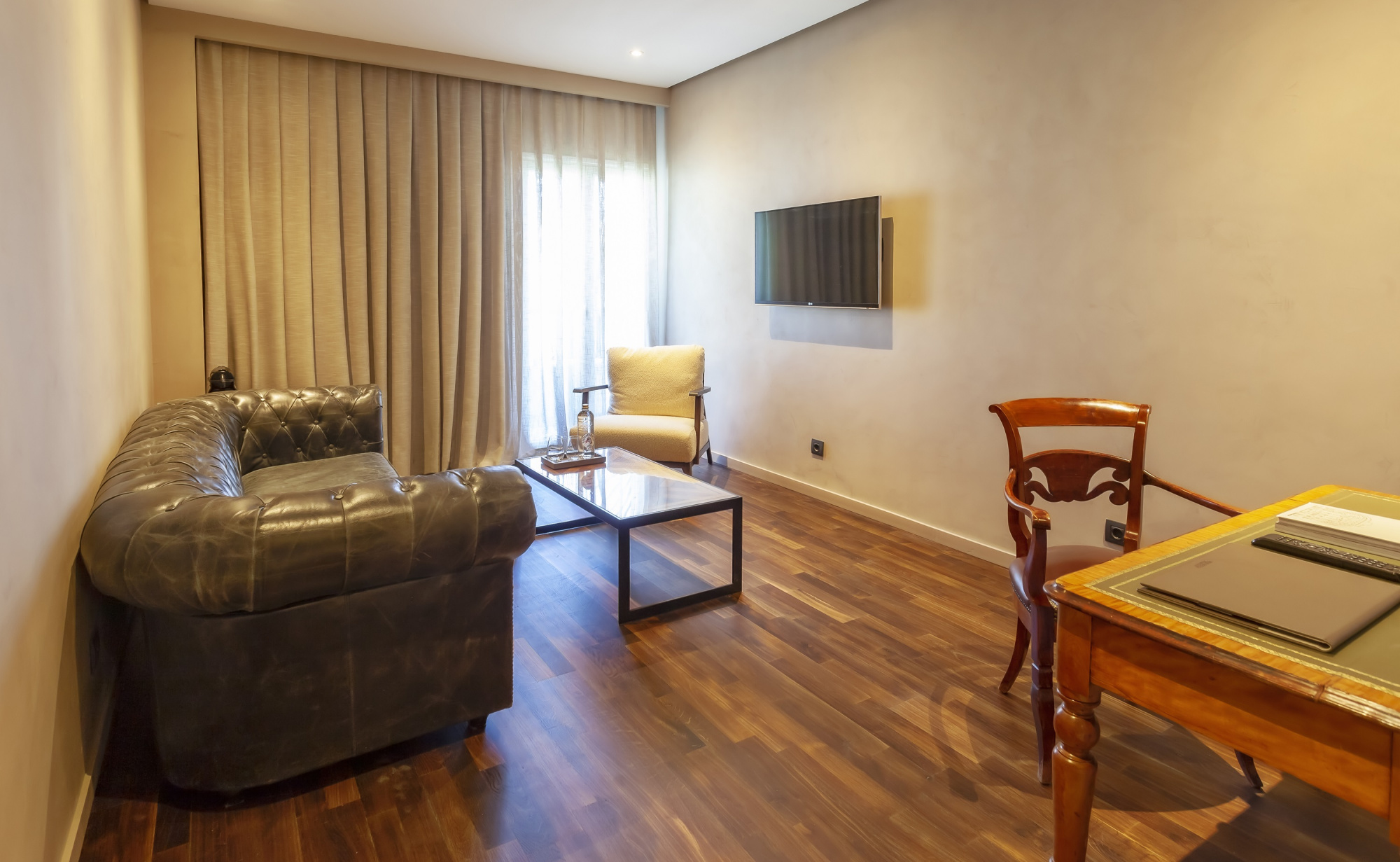La imagen muestra una sala de estar con sofá de cuero, sillón, mesa de centro y cortinas junto a una ventana.