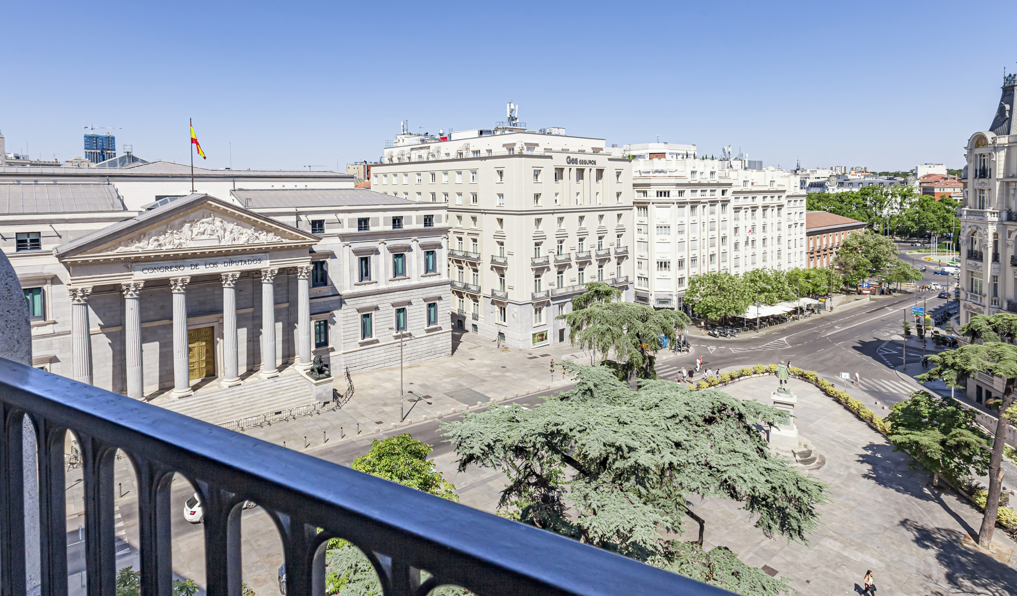 Se ve un balcón con barandilla de metal y una vista de edificios y calles en la ciudad.