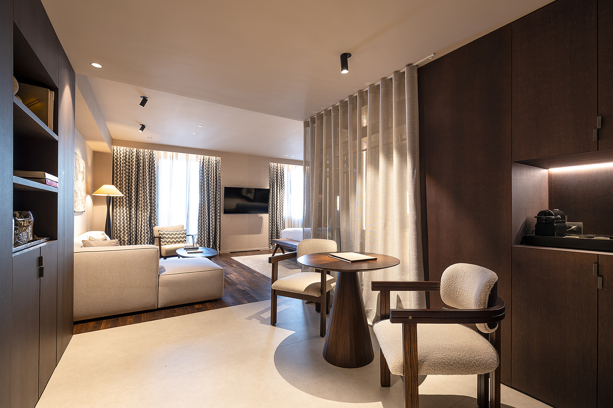 La imagen muestra una suite con cama, zona de estar y una mesa con sillas junto a una ventana con cortinas.