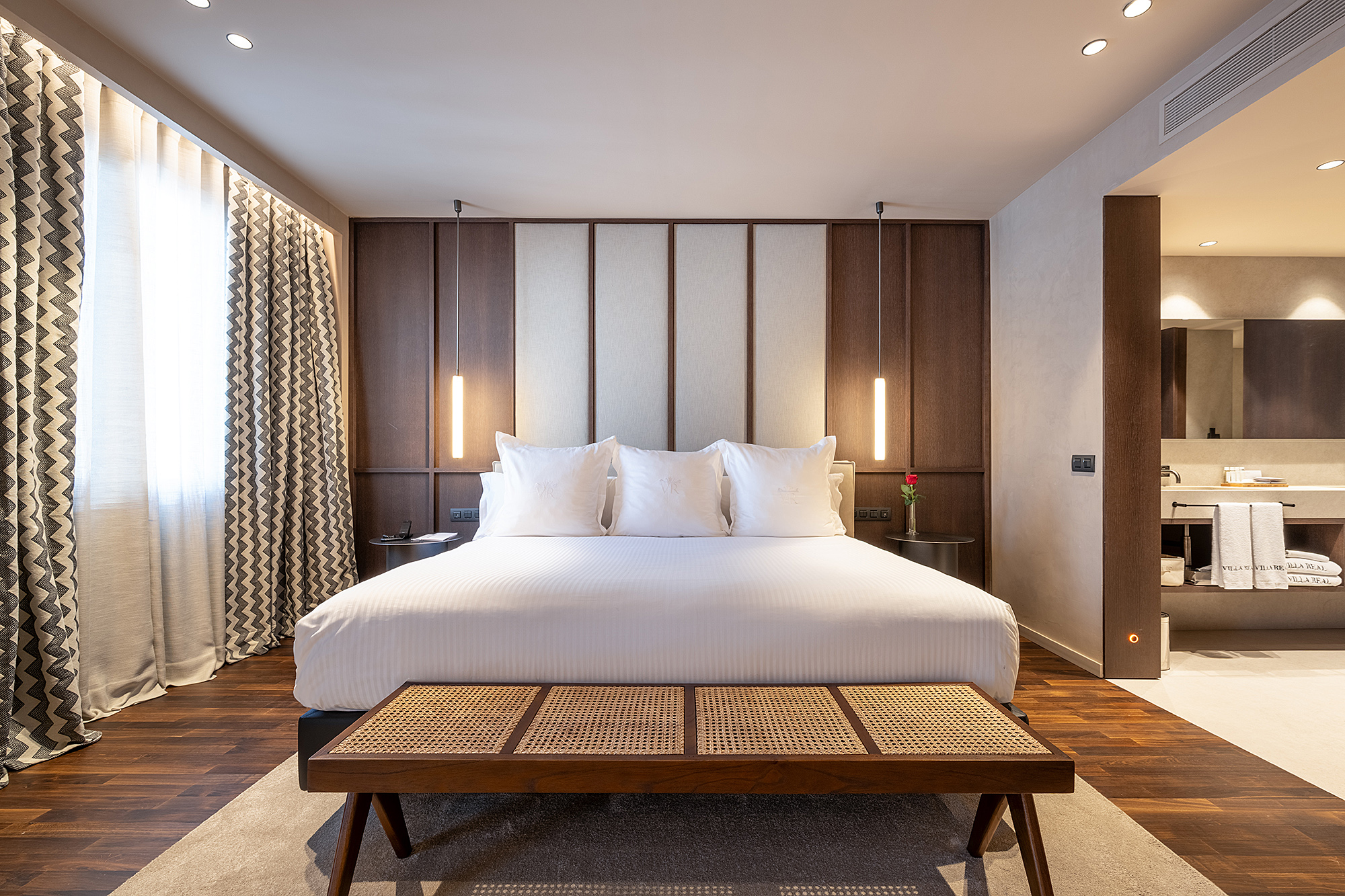 Se ve una habitación con una cama doble, mesitas de noche y cortinas junto a una ventana, con iluminación empotrada en el techo.