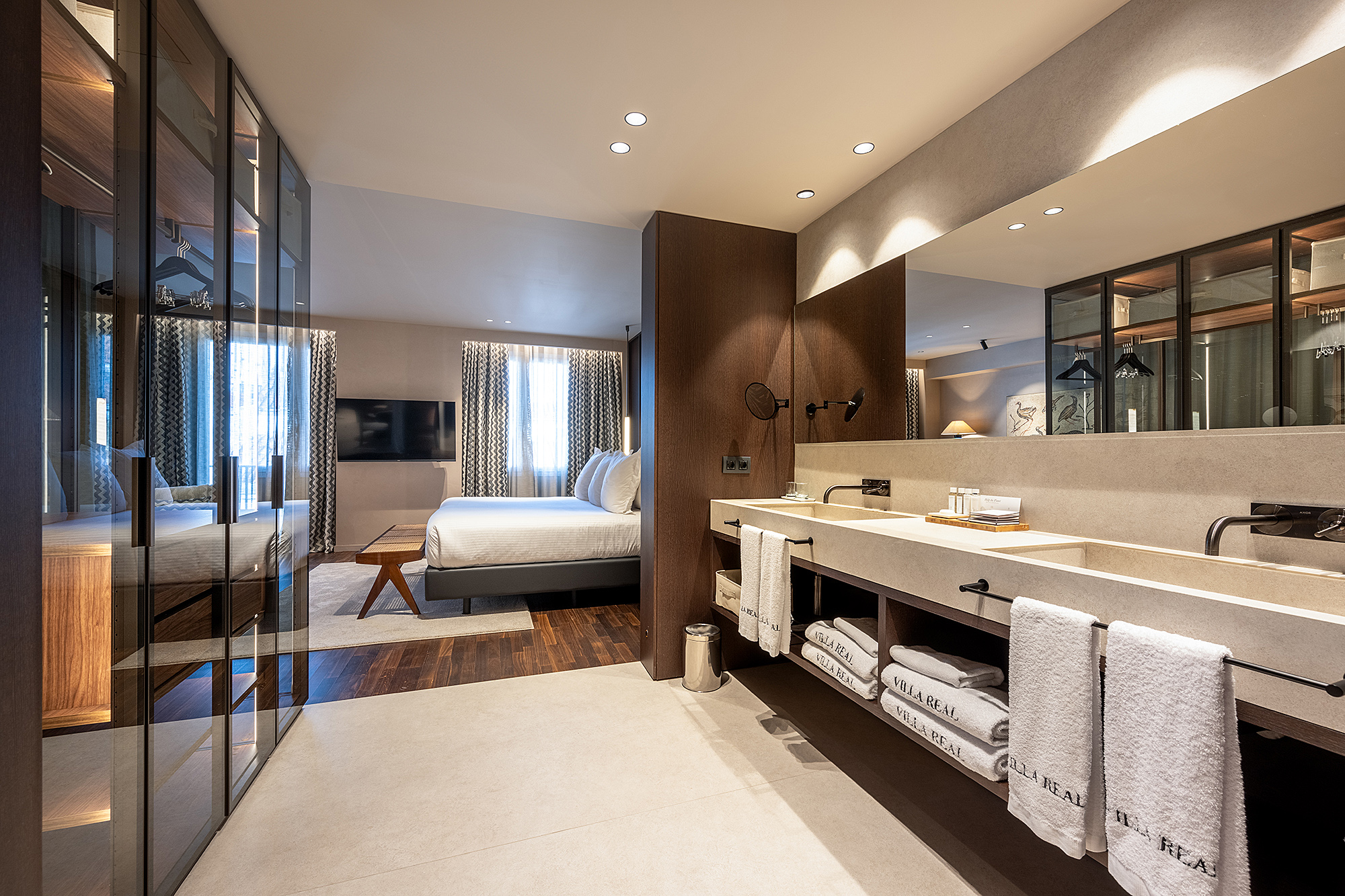 Se ve una suite con cama doble, zona de tocador con lavabo y espejo, y mamparas de vidrio en el baño.