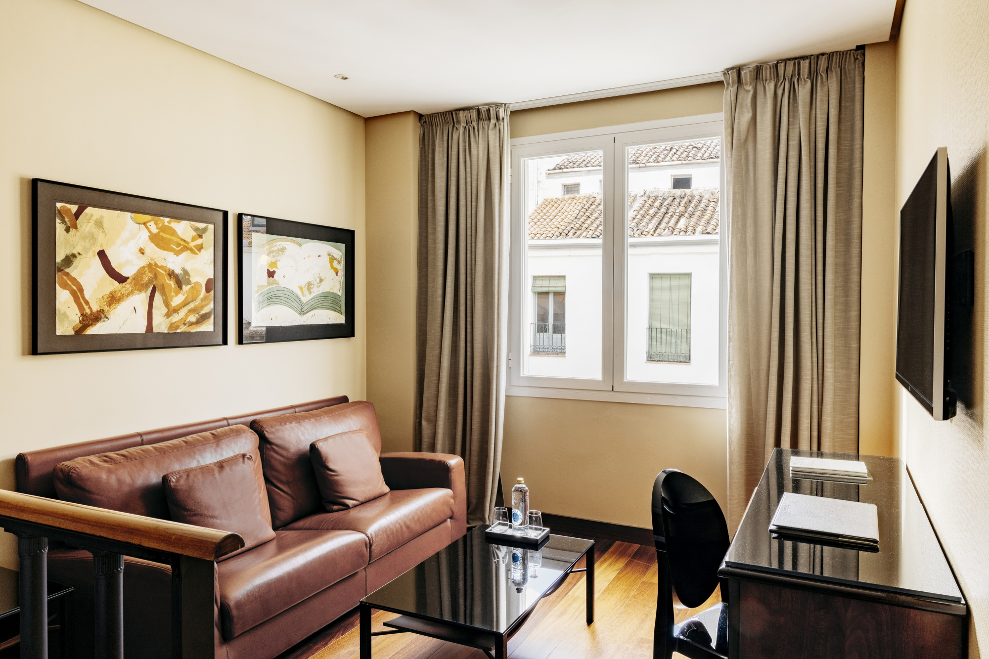 Se ve una sala con sofá de cuero, mesa auxiliar, televisión y una ventana con cortinas.