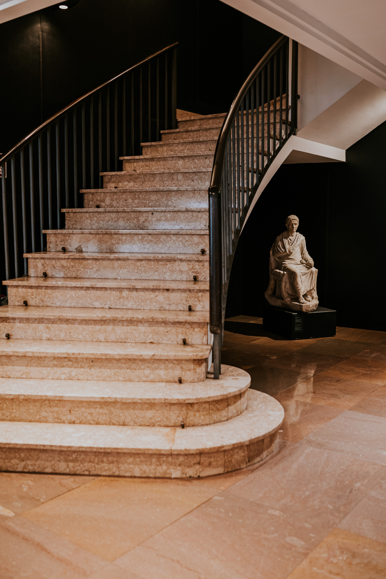 Se ve una escalera de piedra con barandillas negras y una estatua sobre un pedestal en el vestíbulo.