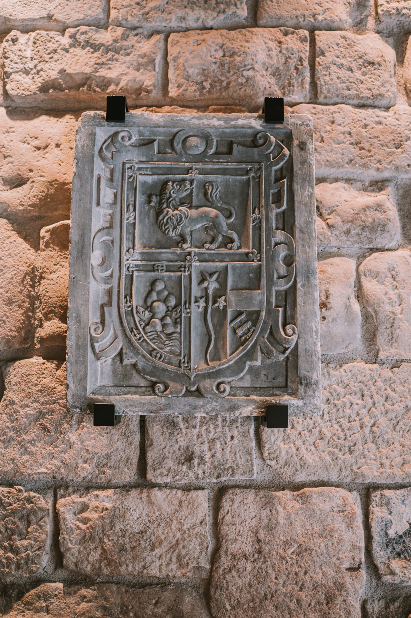 Se ve un escudo decorativo de metal fijado a una pared de piedra con bloques irregulares.
