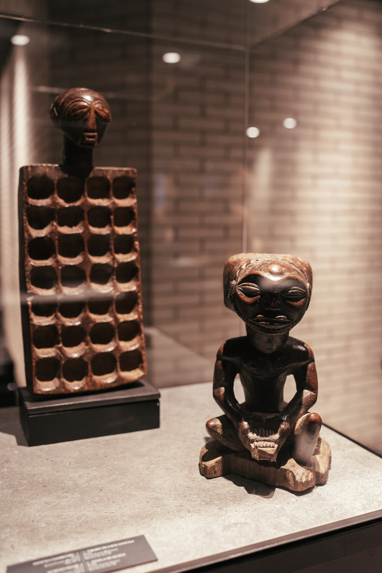 Se ve una escultura africana sobre una mesa y una vitrina con objetos decorativos en una sala con paredes de ladrillo.