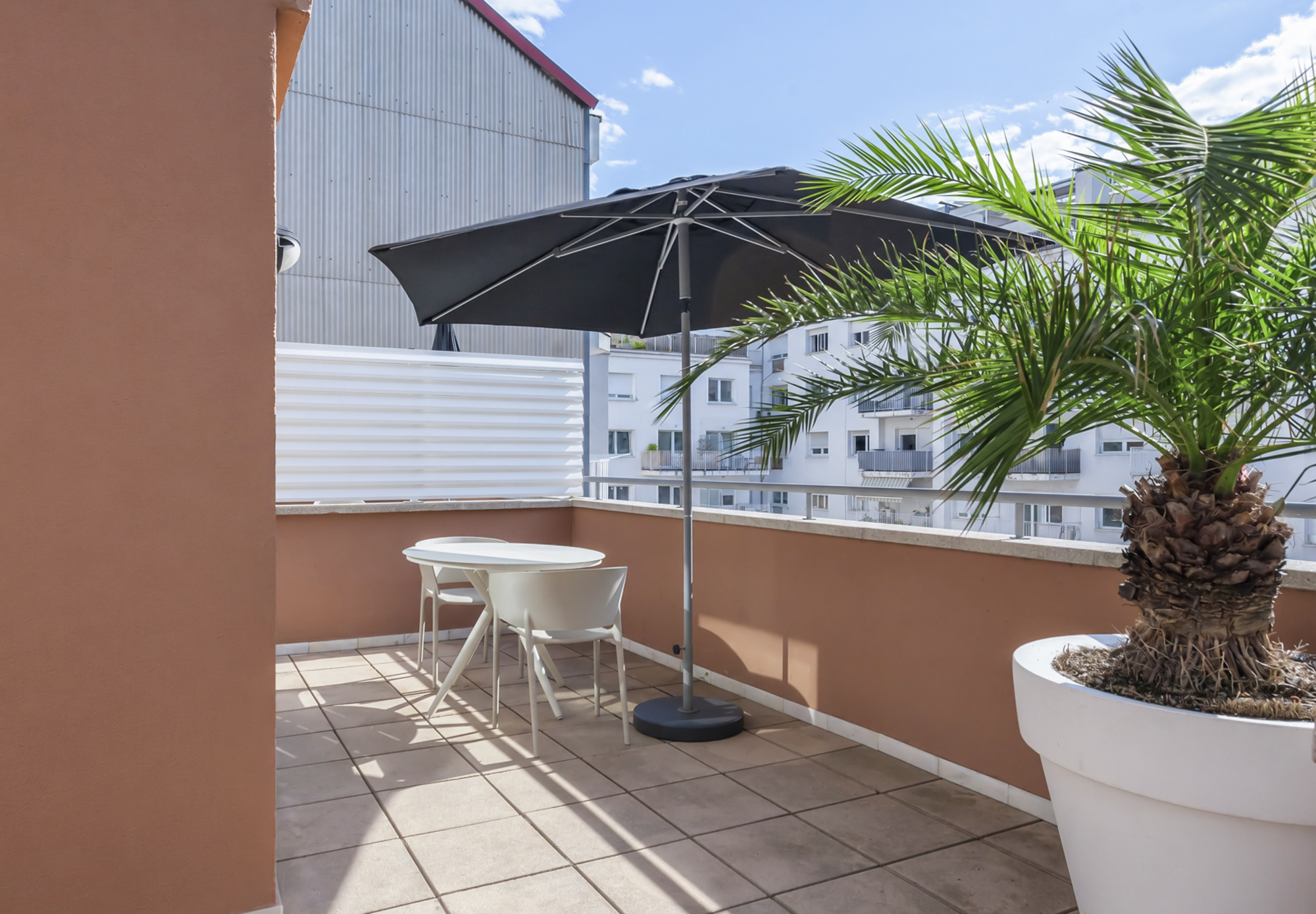 Se ve una terraza exterior con mesa y sombrilla, suelo de baldosas y una jardinera con planta en maceta.