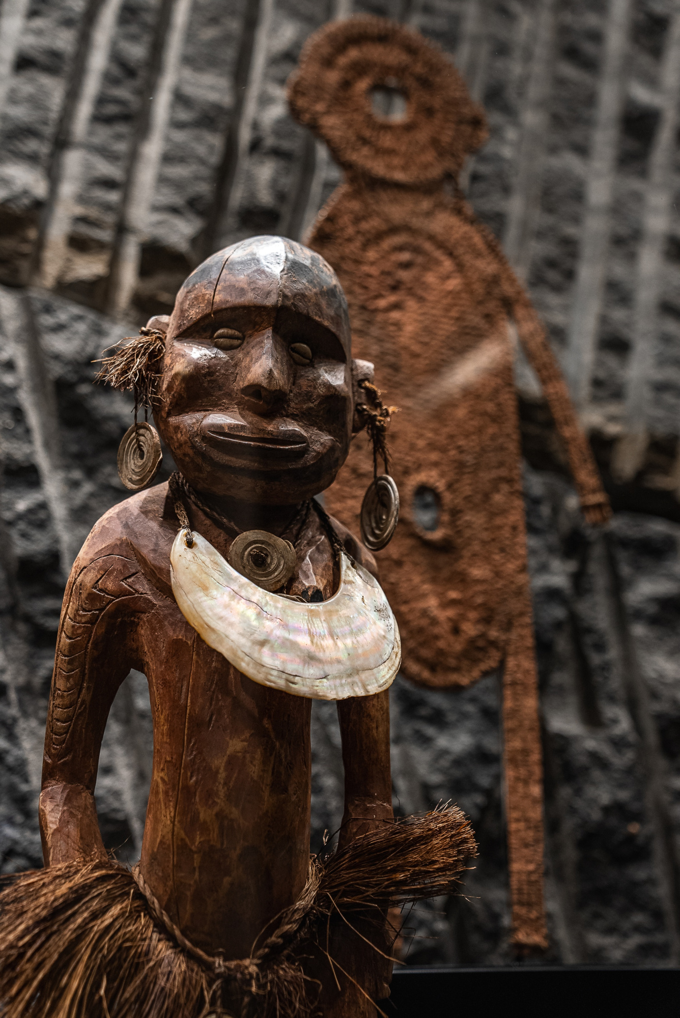 Se ve una escultura decorativa de madera con un tocado y un collar, sobre un fondo con figuras talladas.