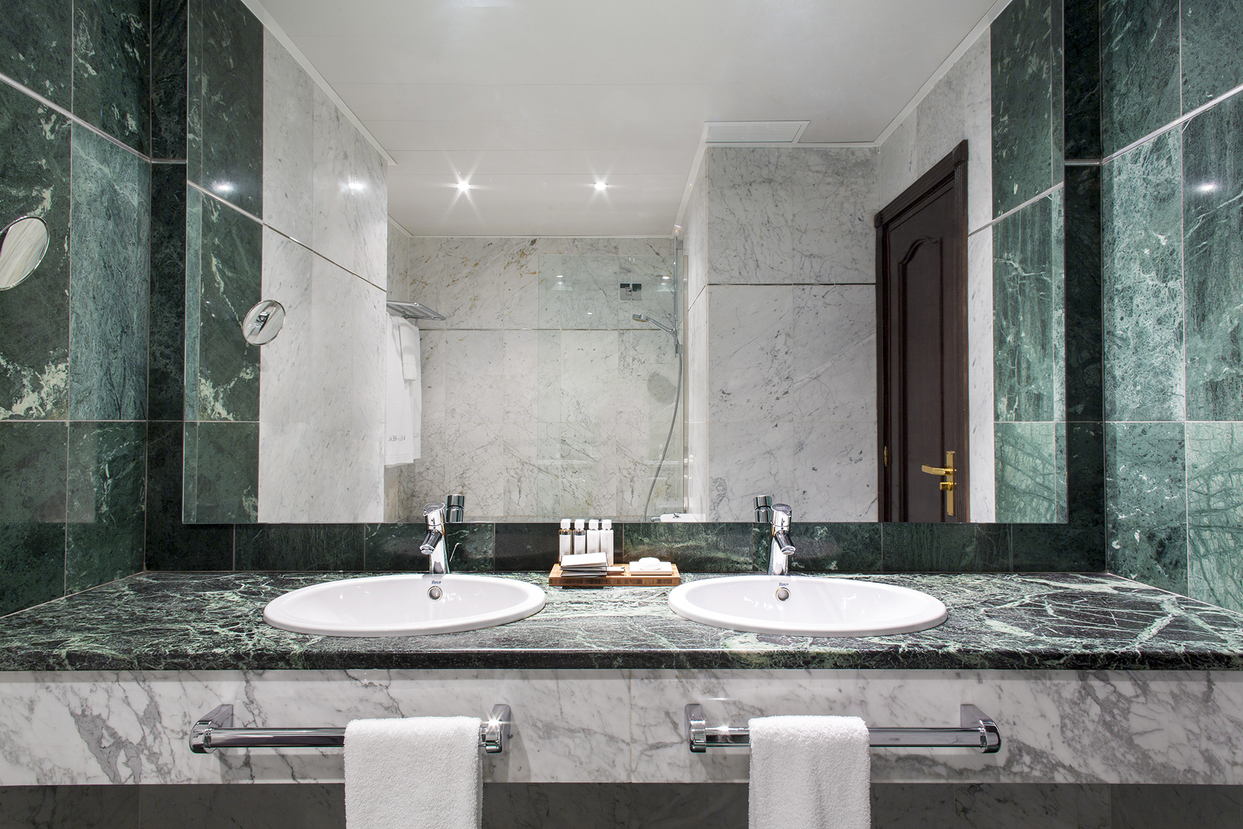 El baño muestra un lavabo doble sobre encimera de mármol, grifería cromada y paredes con azulejos verdes y blancos.