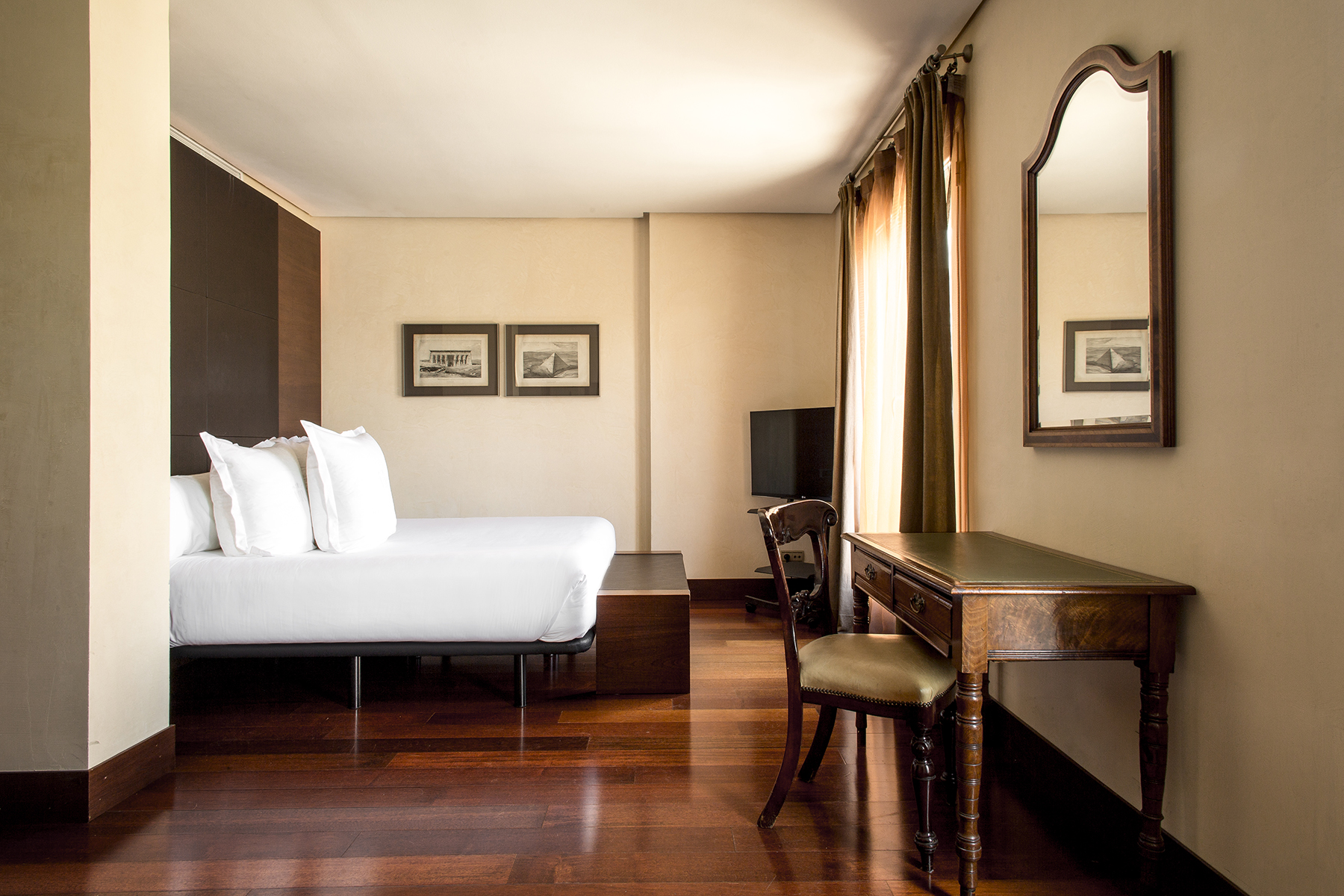 Se ve una cama con ropa de cama blanca, un escritorio con silla y cuadros en las paredes dentro de una habitación.
