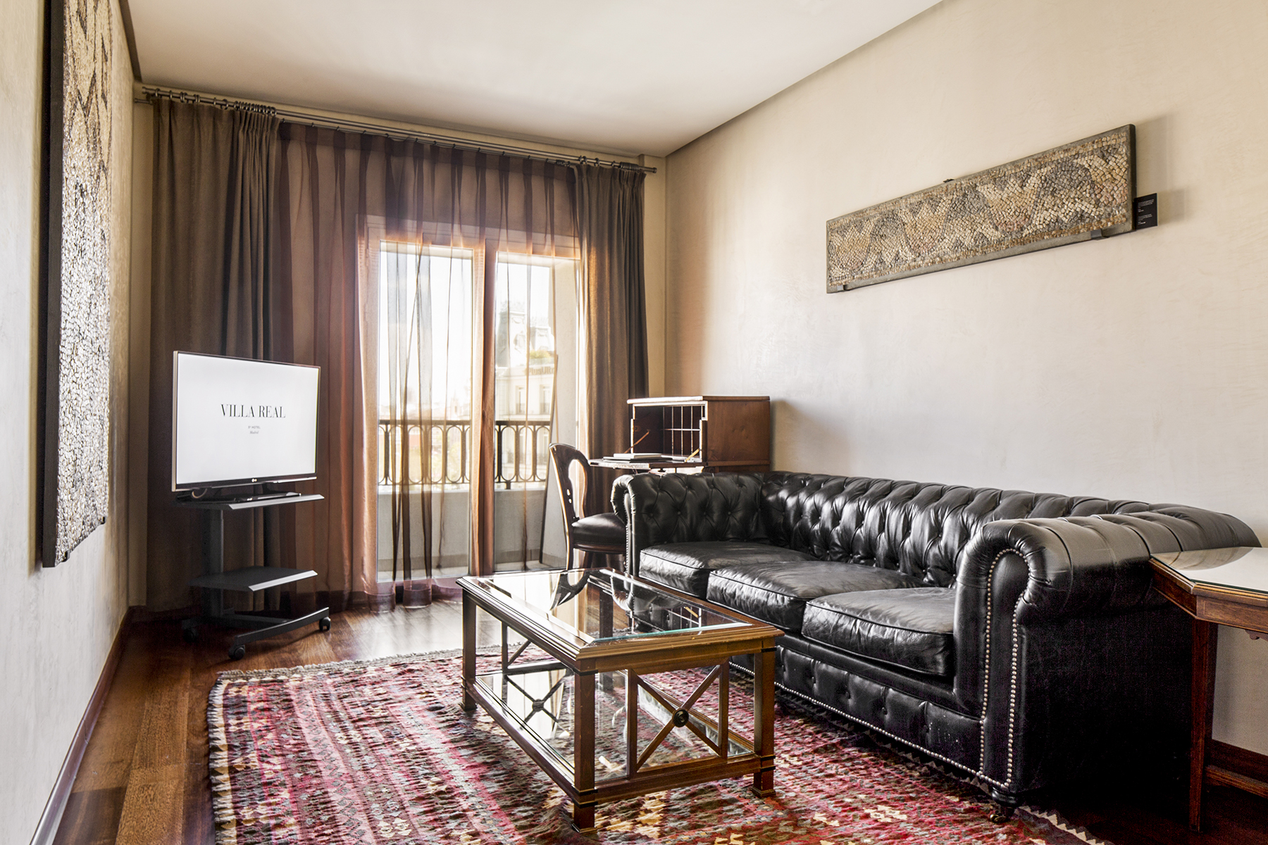 La imagen muestra una sala de estar con sofá de cuero, mesa de centro, TV y cortinas junto a una puerta con balcón.