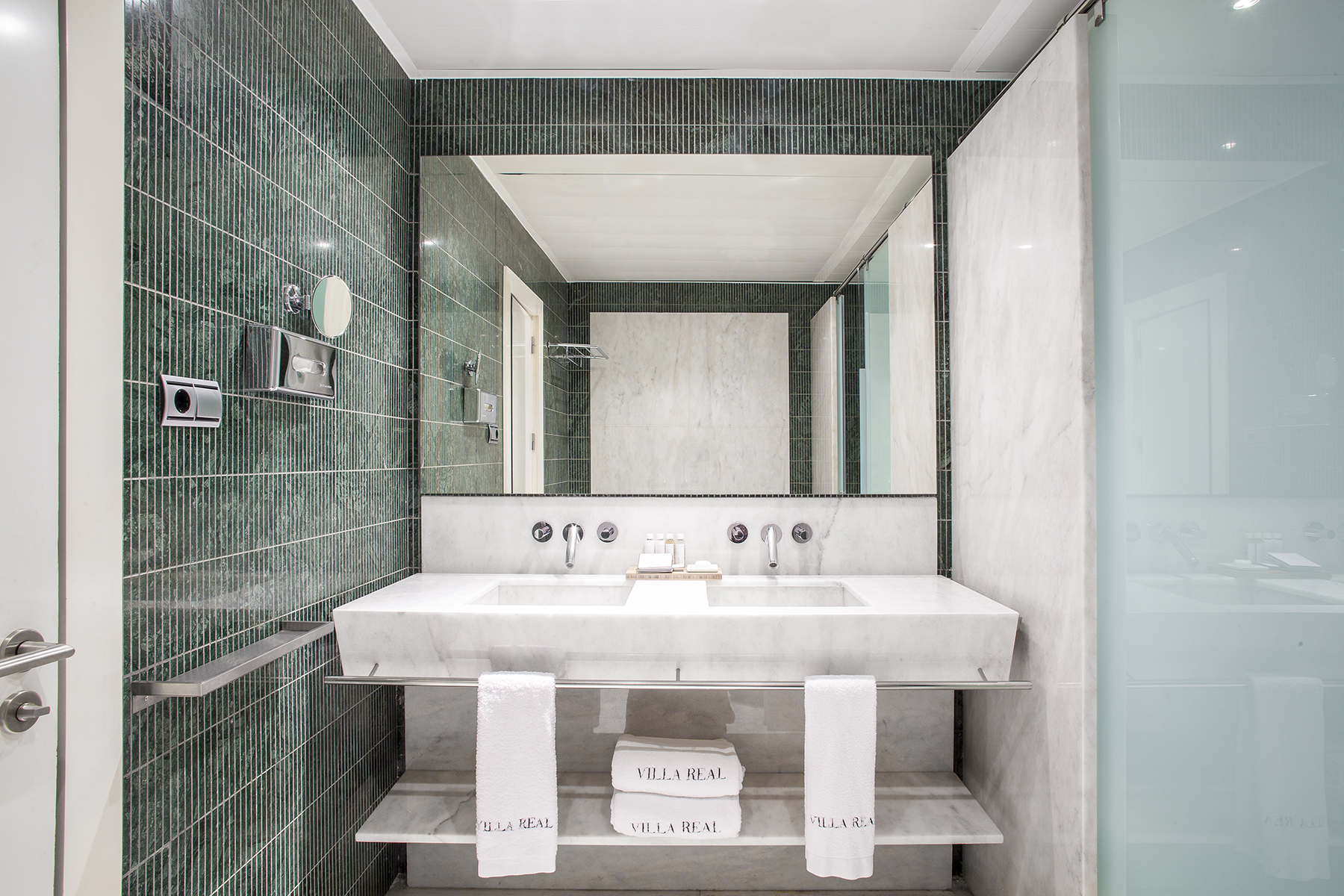 El baño muestra un doble lavabo sobre una encimera blanca, espejos y una ducha con mampara de vidrio.
