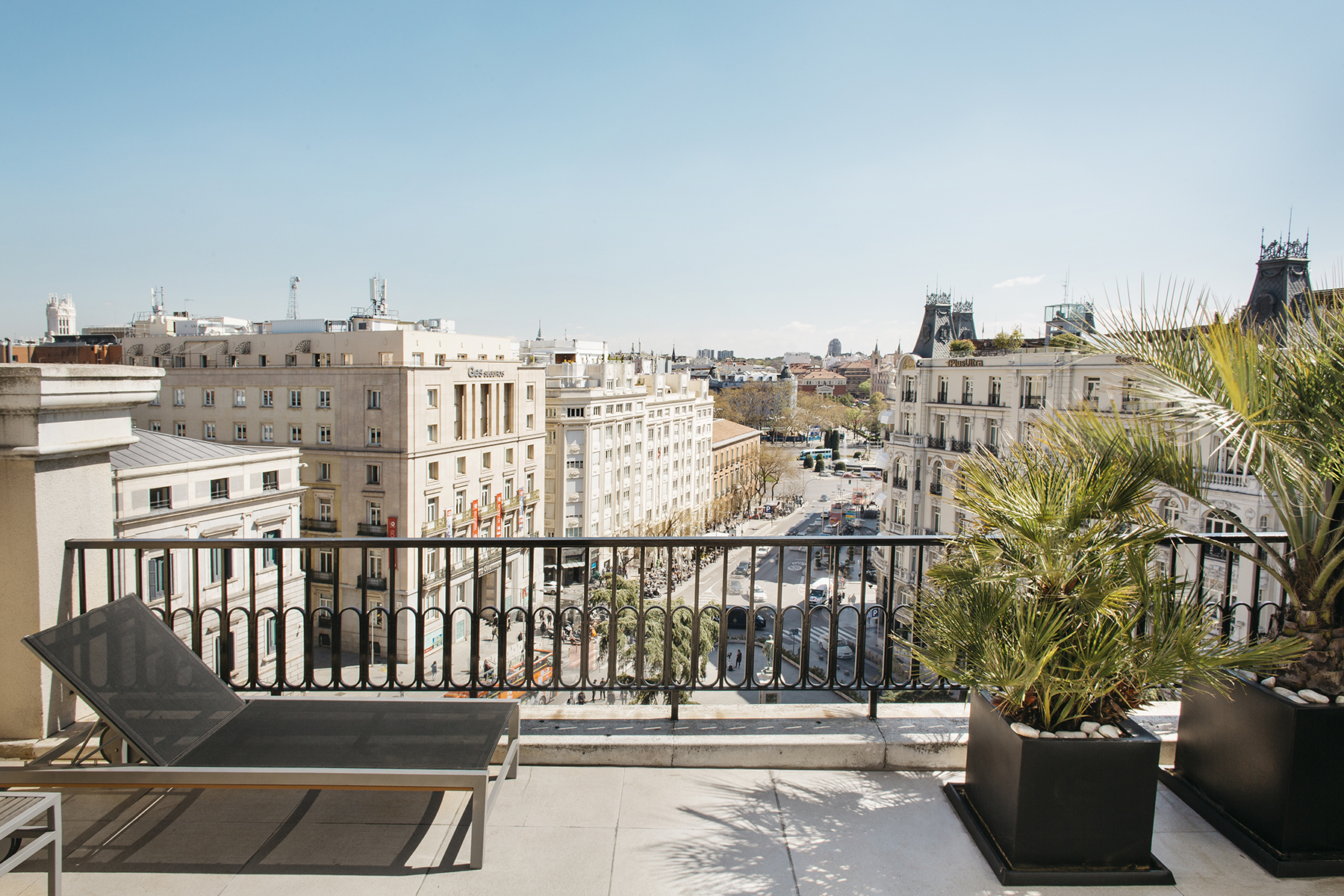 Se ve una terraza con suelo claro, barandilla de metal y asientos junto a plantas en macetas, con edificios al fondo.