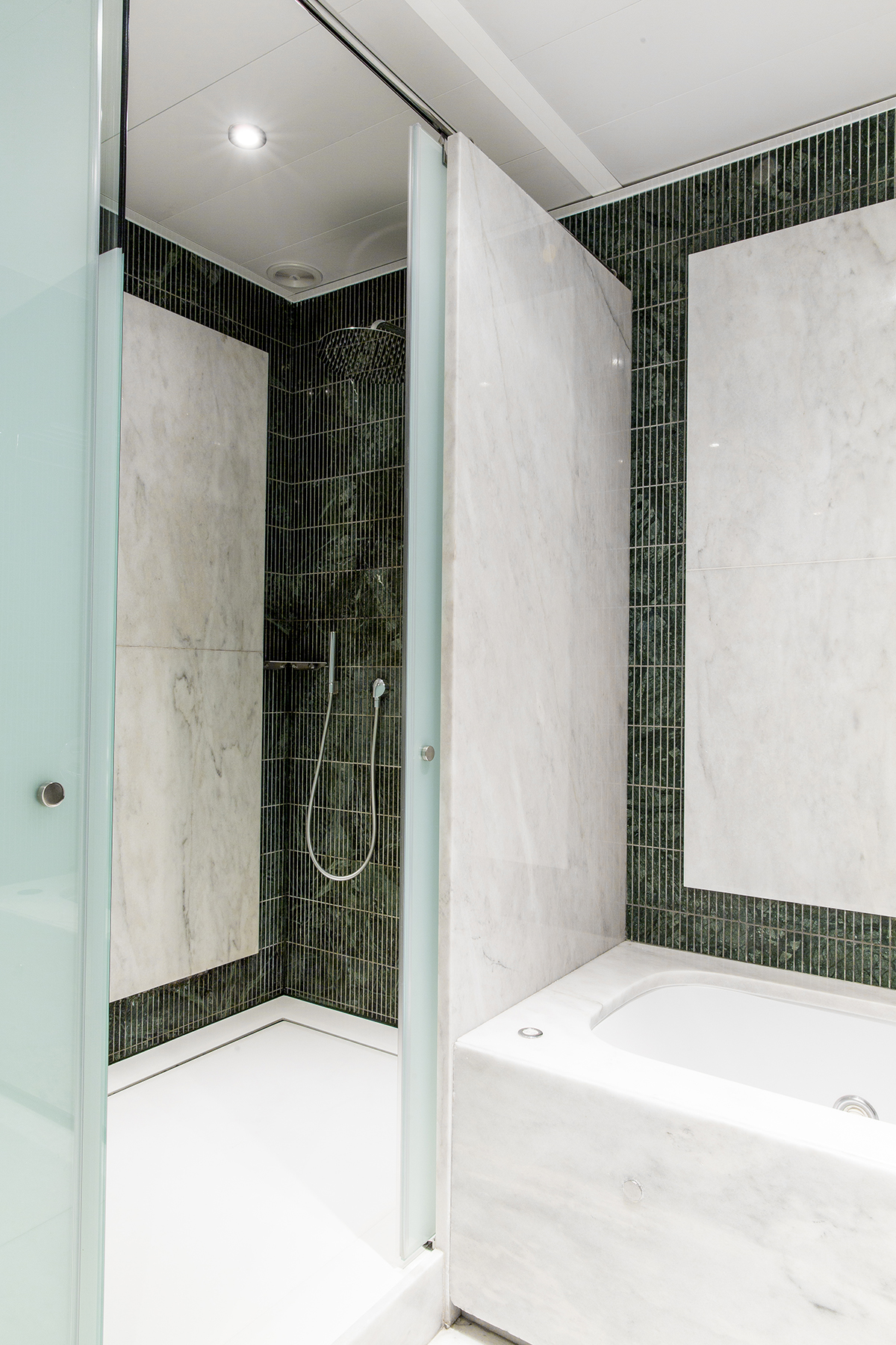 Se ve una ducha con mampara de vidrio y una bañera blanca en un baño con paredes revestidas de azulejos oscuros y claros.
