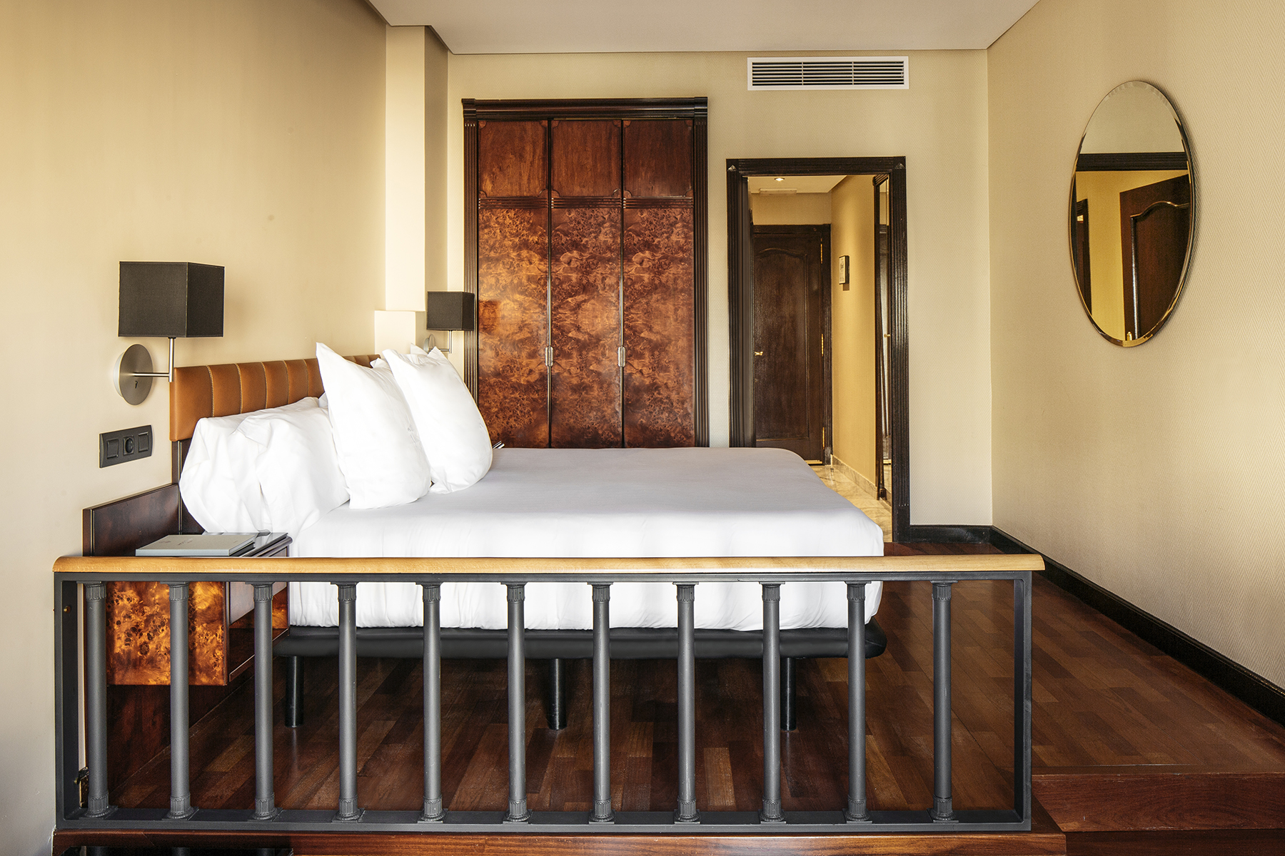 Se ve una cama doble con cabecero de madera, mesita y lámpara, junto a una pared con puerta y un espejo redondo.