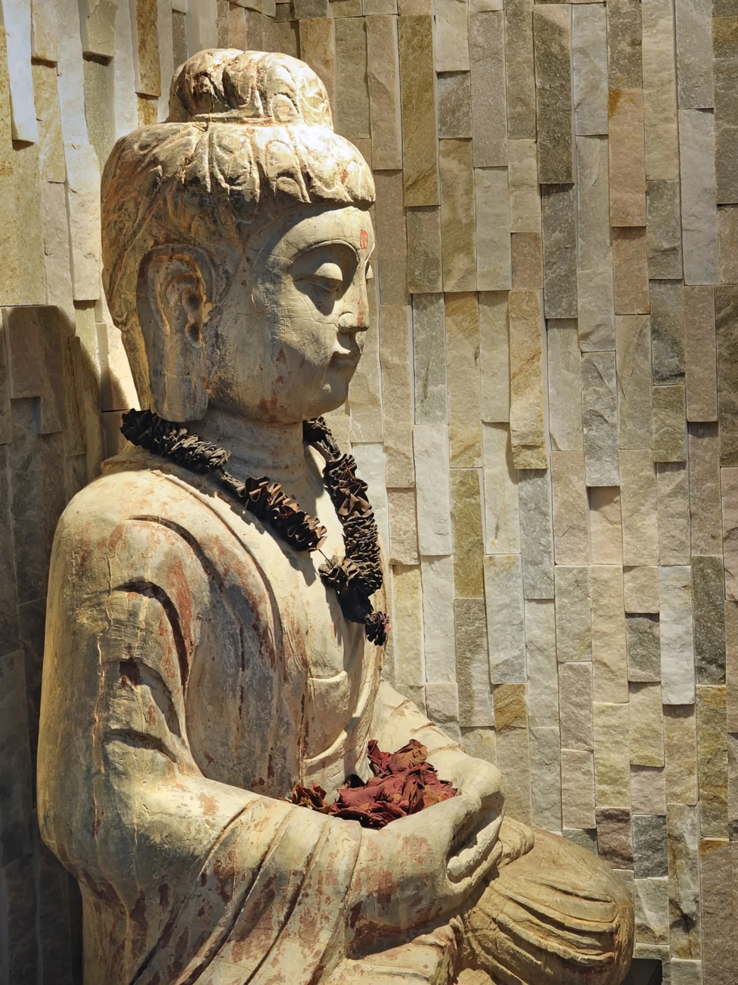 Una escultura de piedra con las manos abiertas aparece junto a un muro con relieve de bloques de piedra.