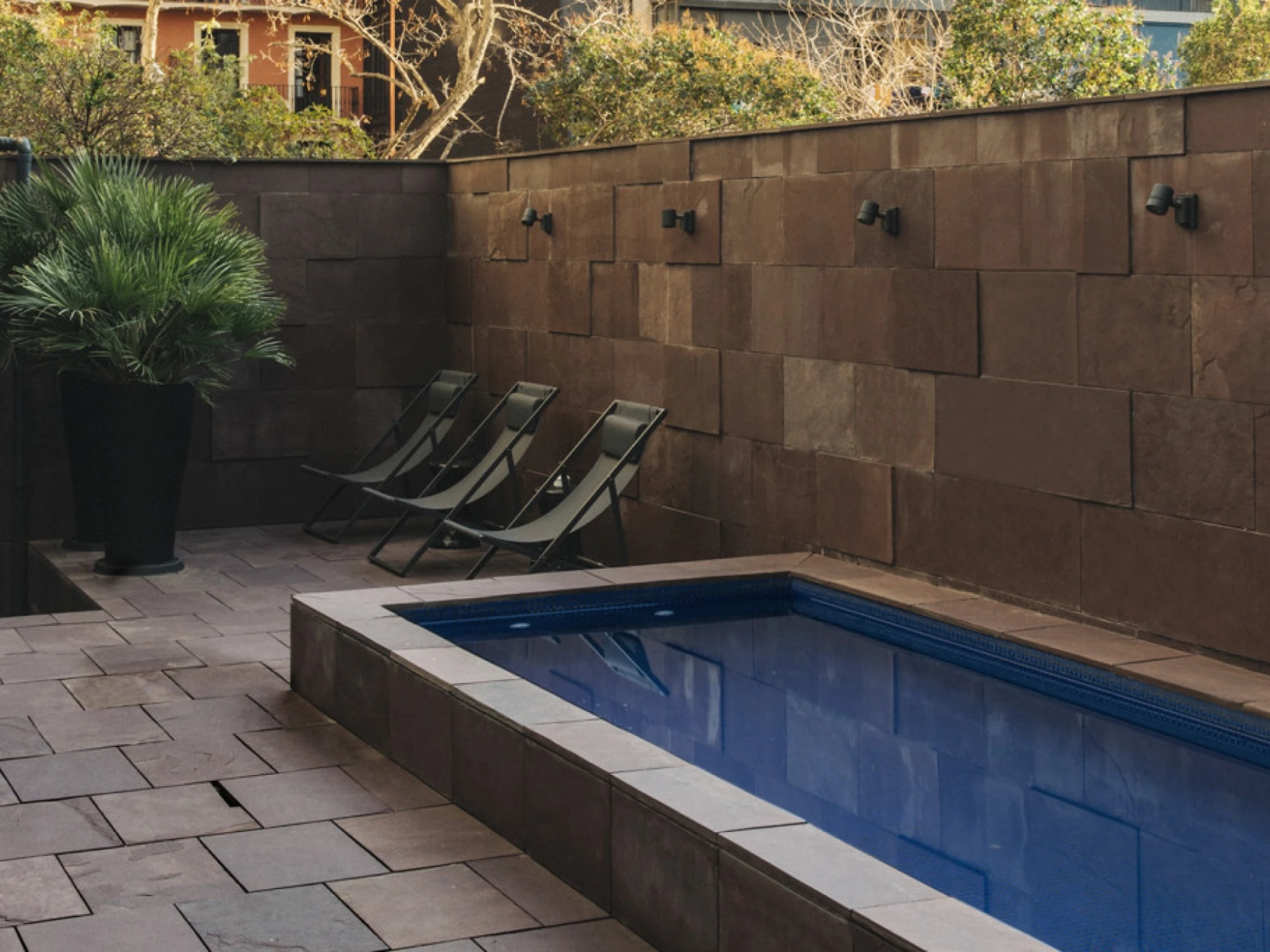 Se ve una piscina rectangular exterior con bordes oscuros y varias tumbonas en un patio con suelo de losas.